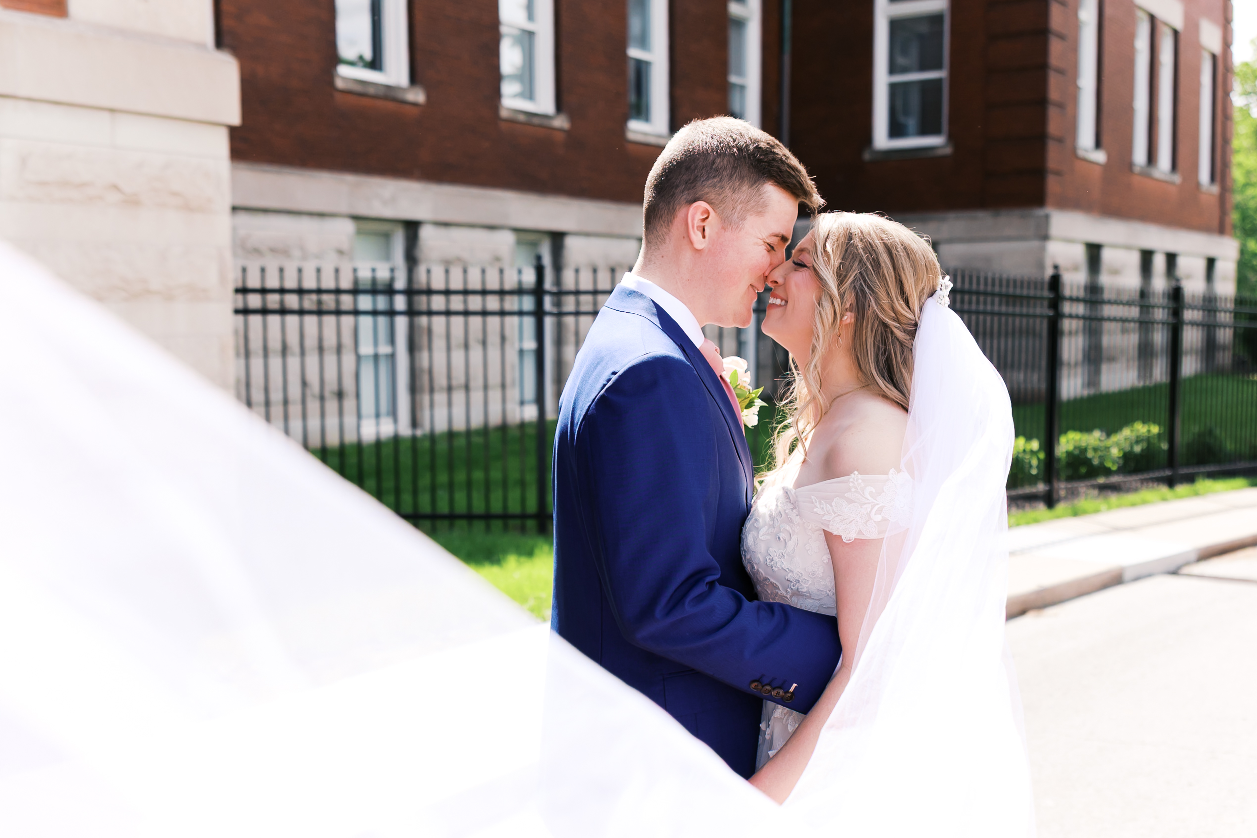 Bride and groom smile forehead to forehead outdoors with soft sunlight and flowing veil during wedding portraits.
