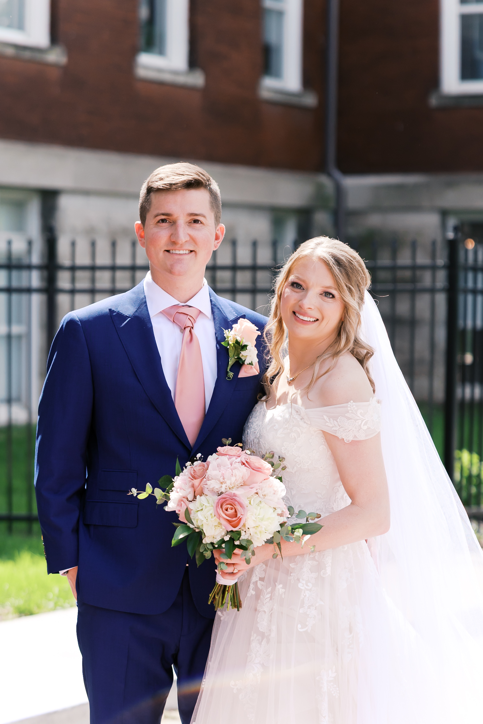 Bride and groom smile at the camera while holding a blush and white floral bouquet outside wedding venue.