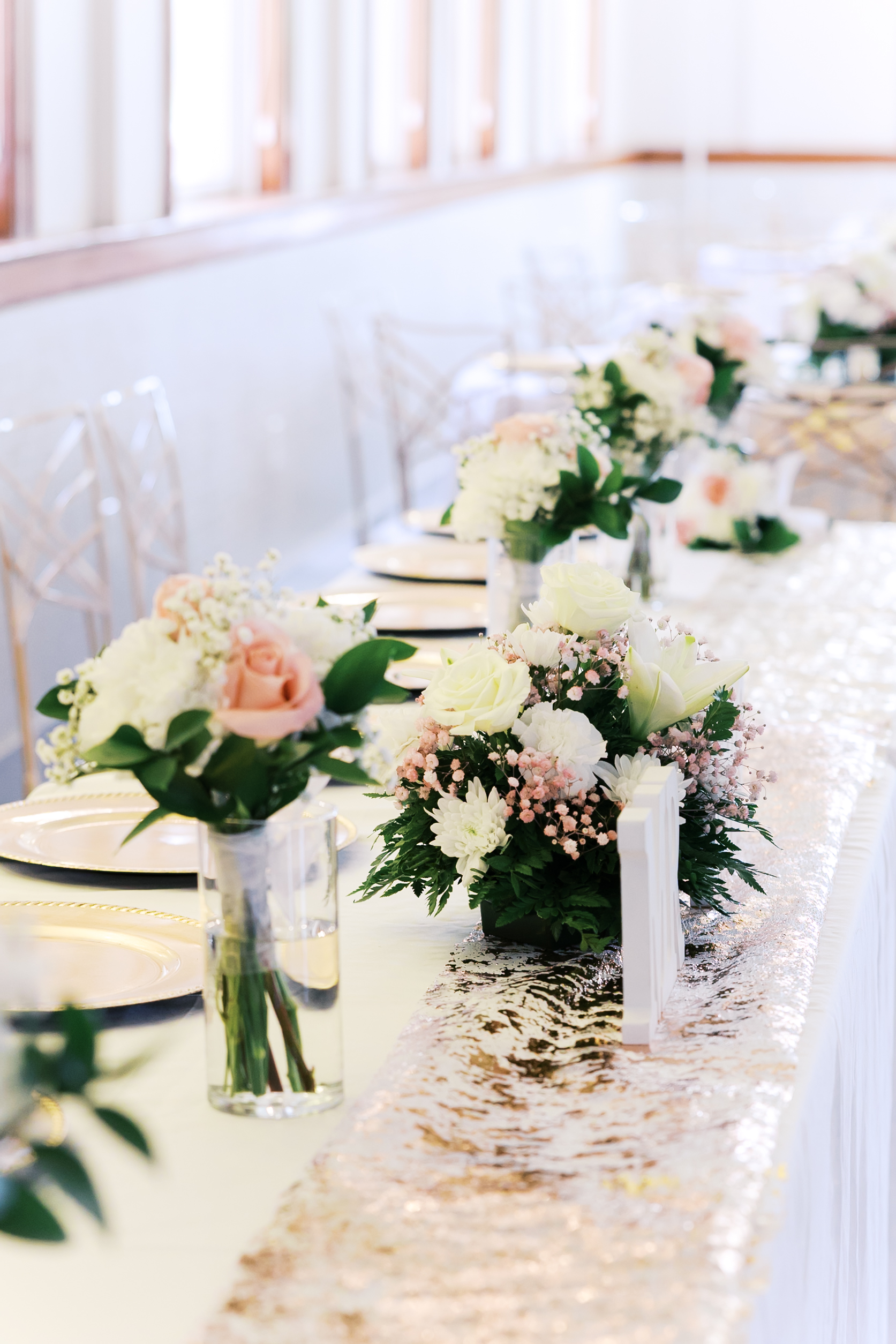 Wedding reception head table with blush and white floral centerpieces, elegant place settings, and shimmering runner.