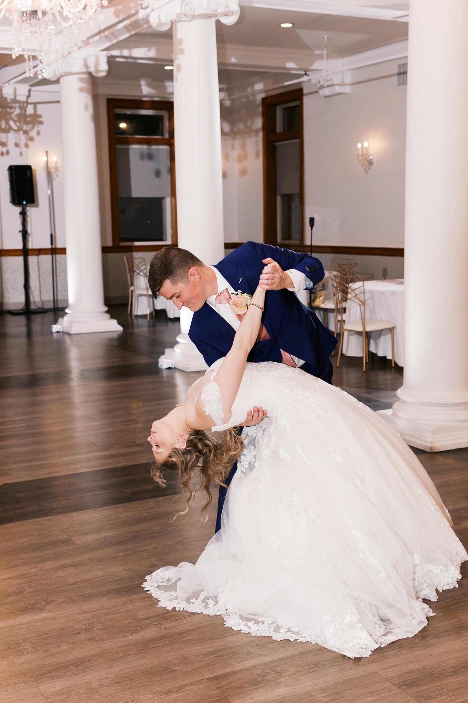 Bride and groom perform a dramatic first dance dip in an elegant ballroom with white columns and chandeliers.