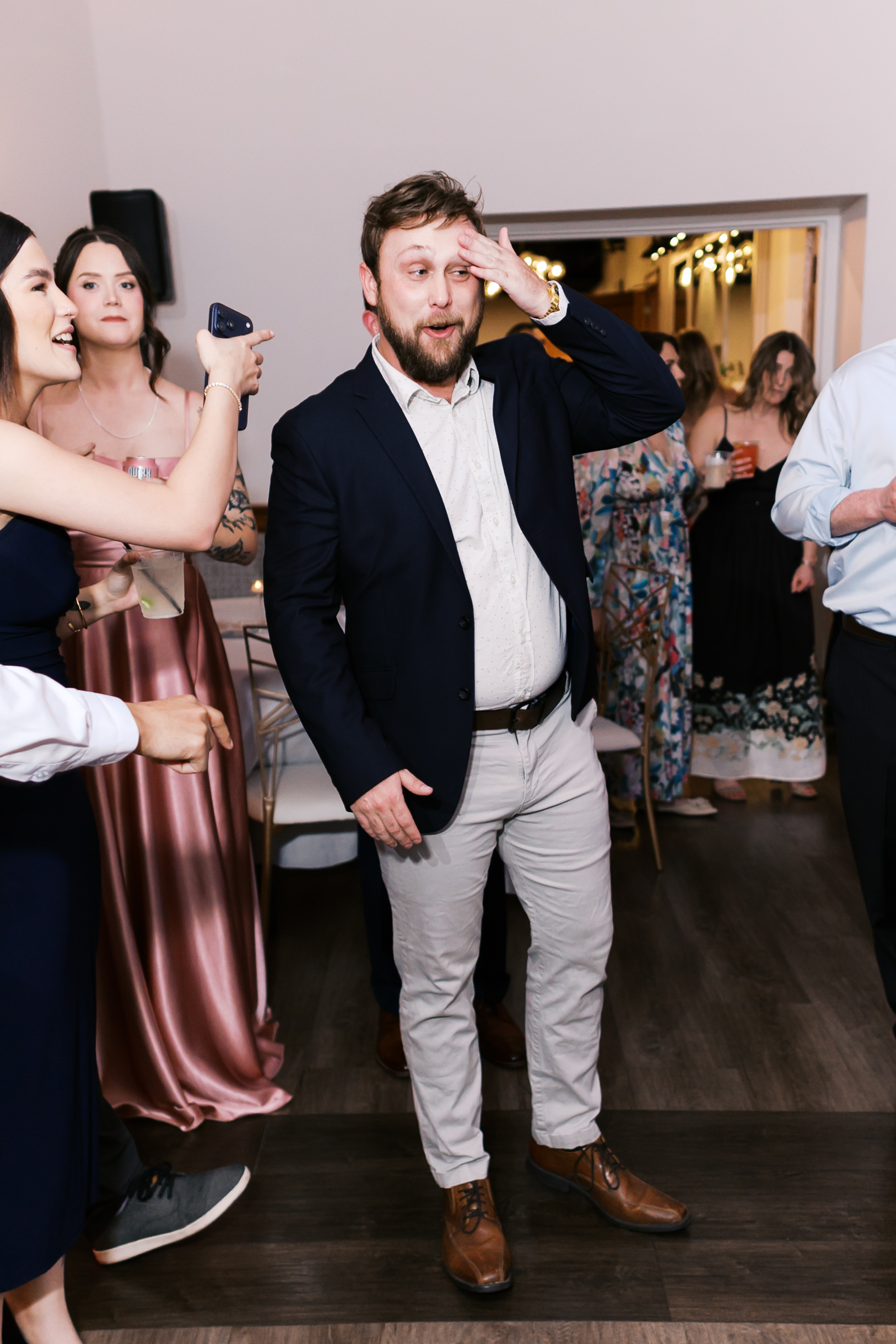 Wedding guest reacts during bouquet toss with hand on forehead as other guests laugh and watch on the reception dance floor.