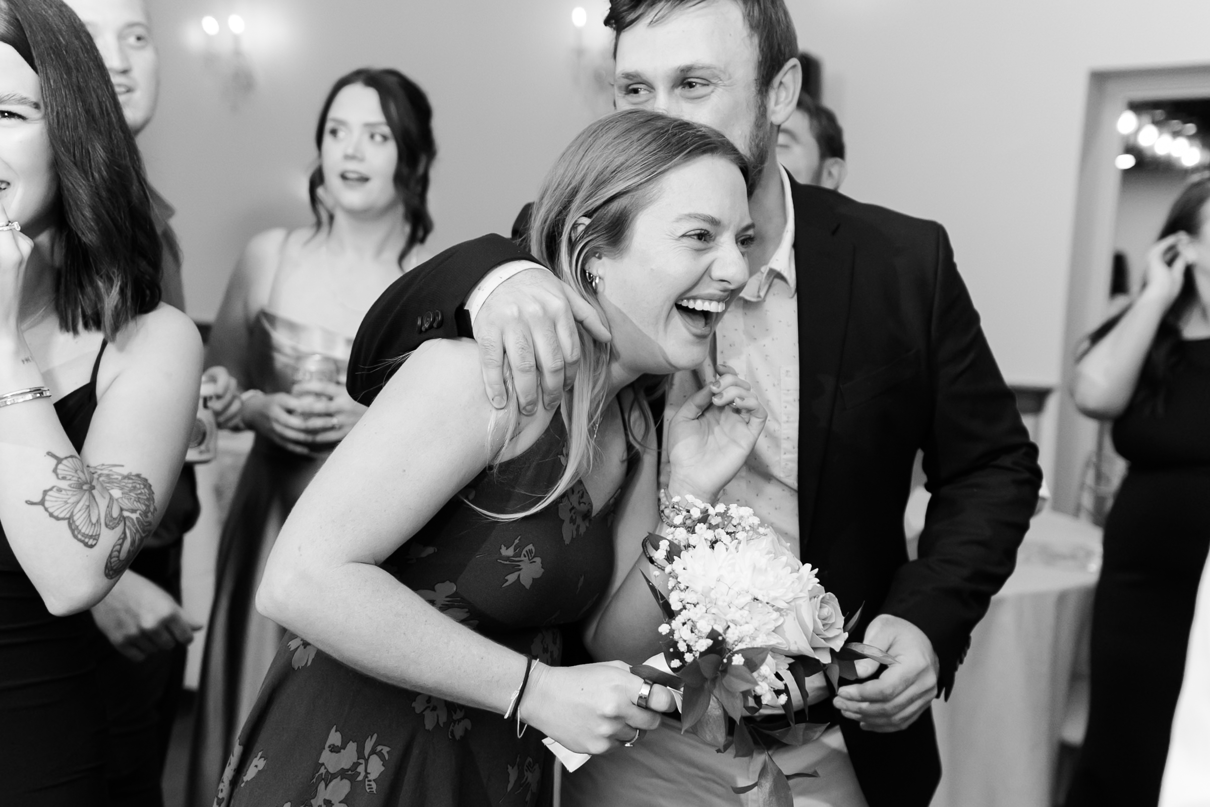 Wedding guest hugs a smiling woman holding the bouquet after the bouquet toss while friends laugh at the reception.