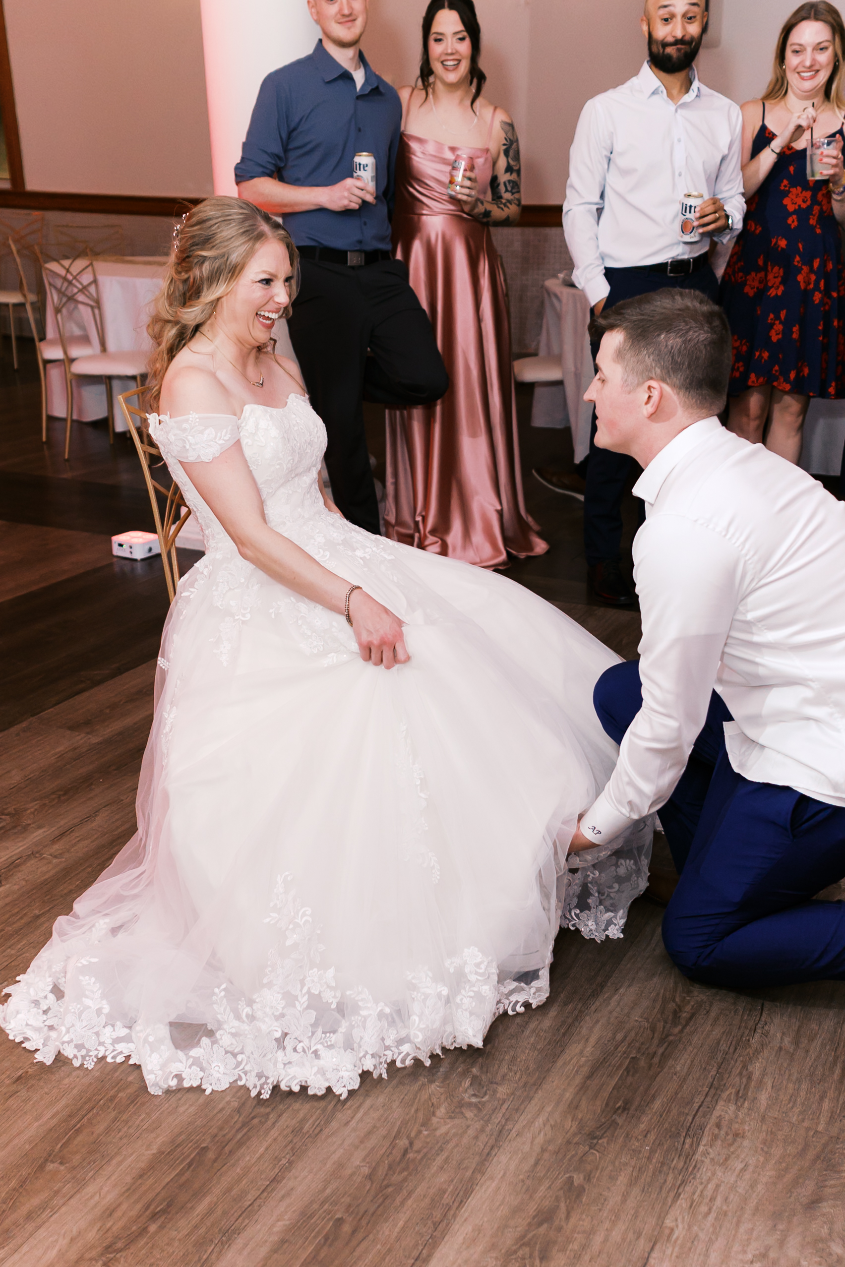 Bride laughs as groom kneels to remove her garter while wedding guests watch and smile during the reception.