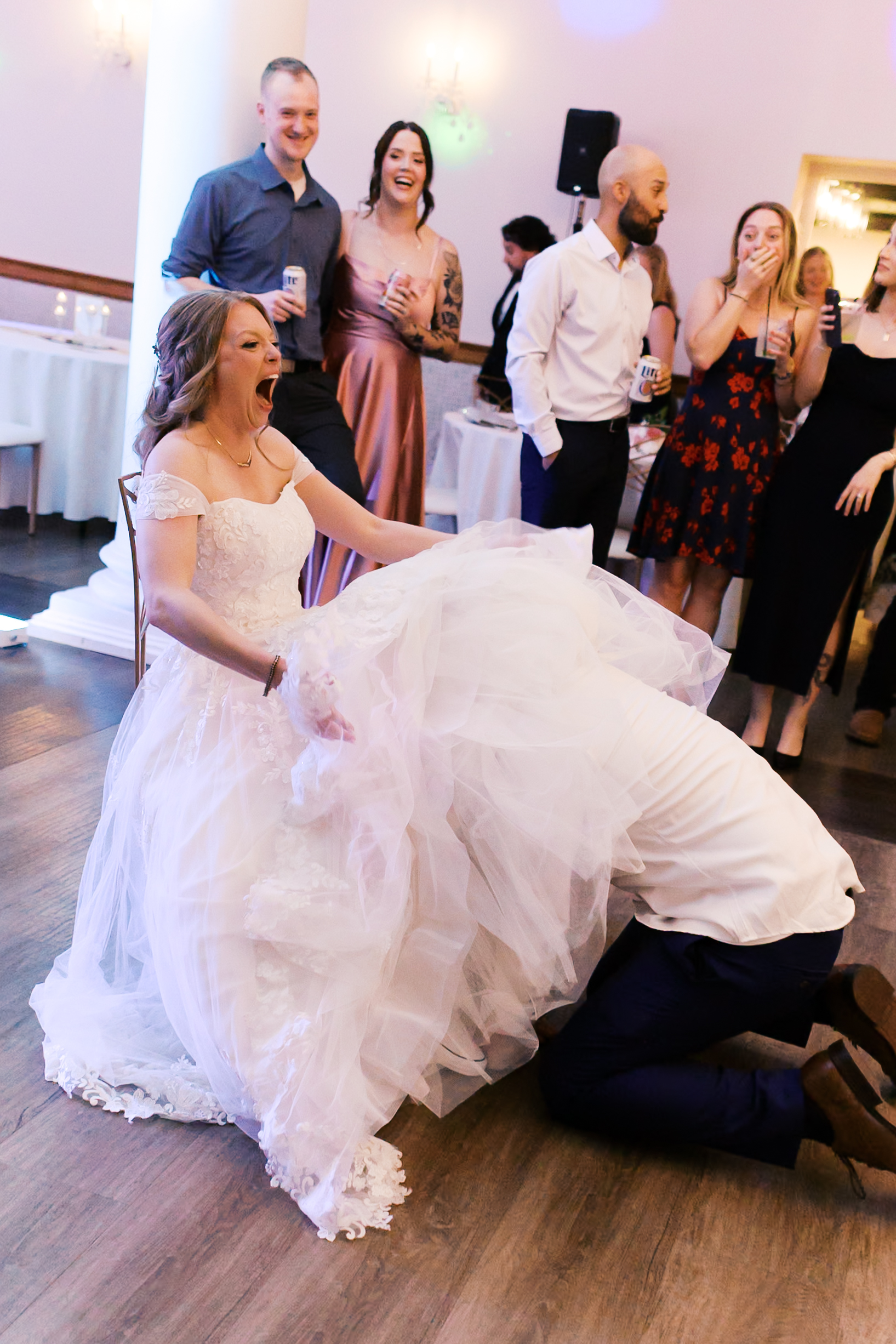 Bride laughs with her wedding dress lifted as groom ducks underneath to retrieve the garter while guests react around them.