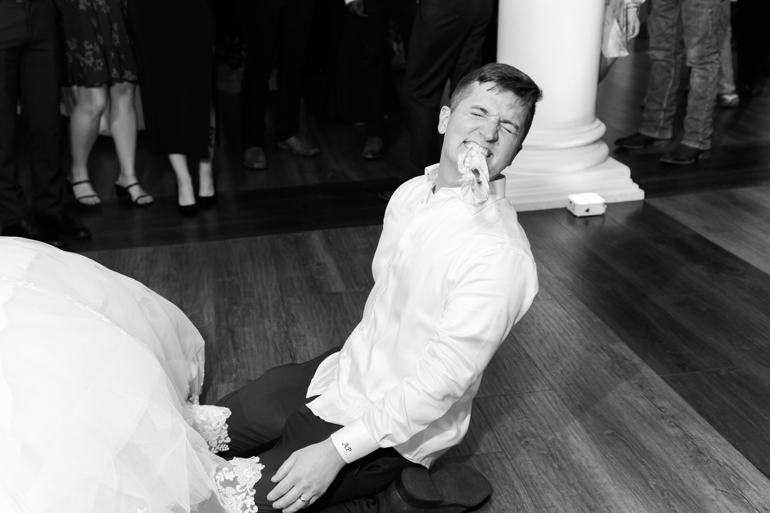 Groom sits on the dance floor holding the bridal garter in his teeth after the garter toss as guests look on.