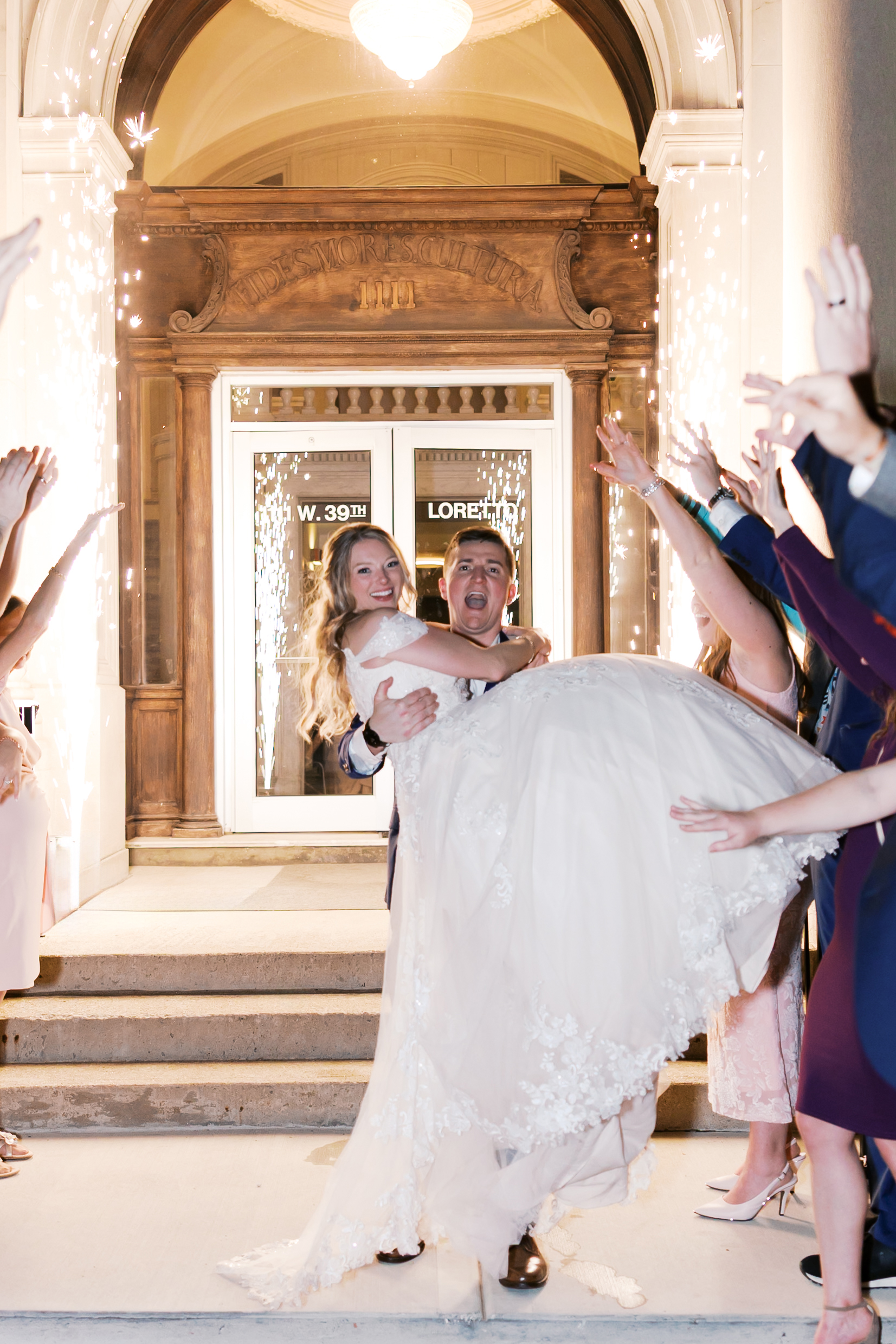 Groom carries the bride through a sparkler exit as wedding guests cheer and celebrate outside the reception venue.