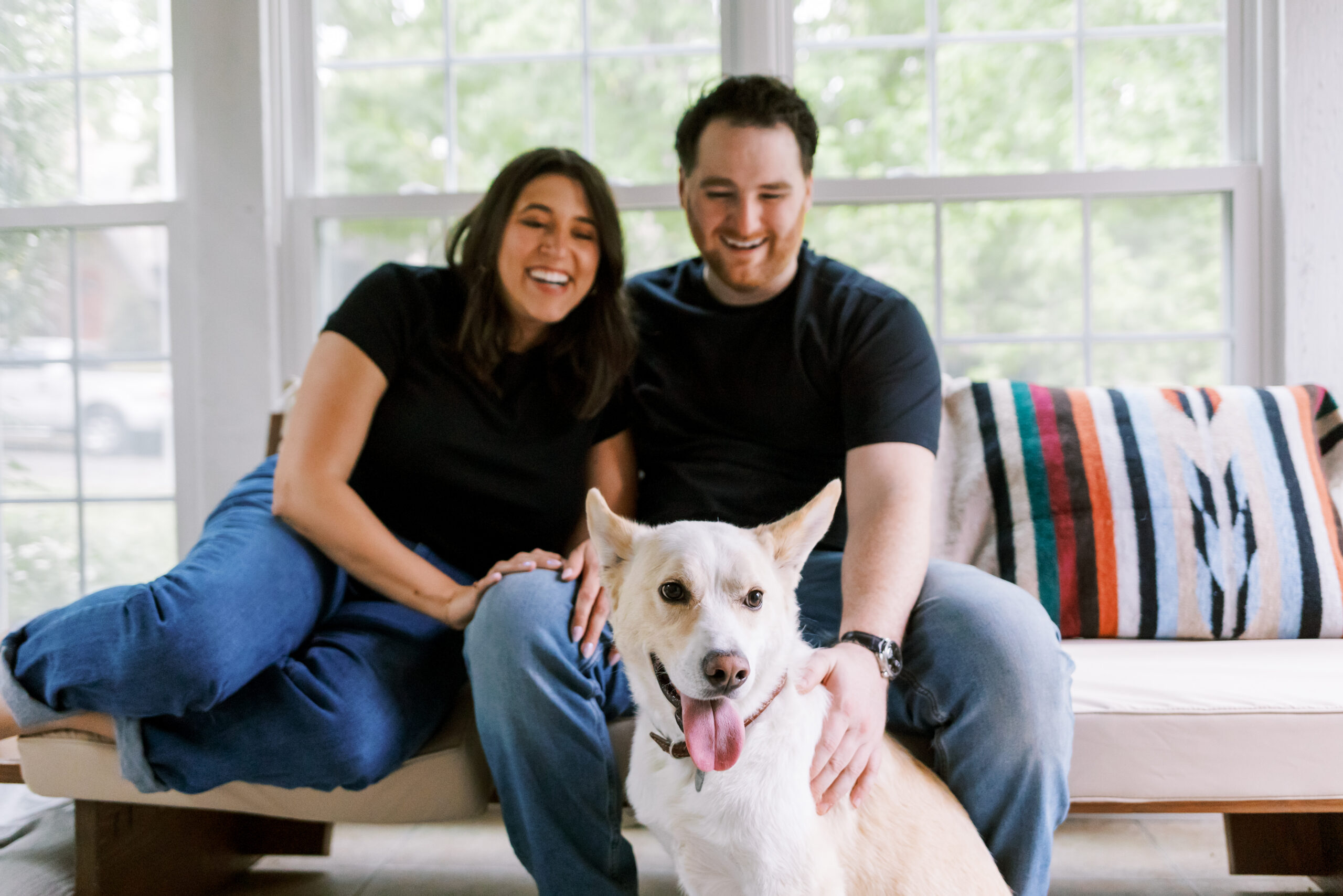 Engaged couple laughing on a couch with their dog at home.