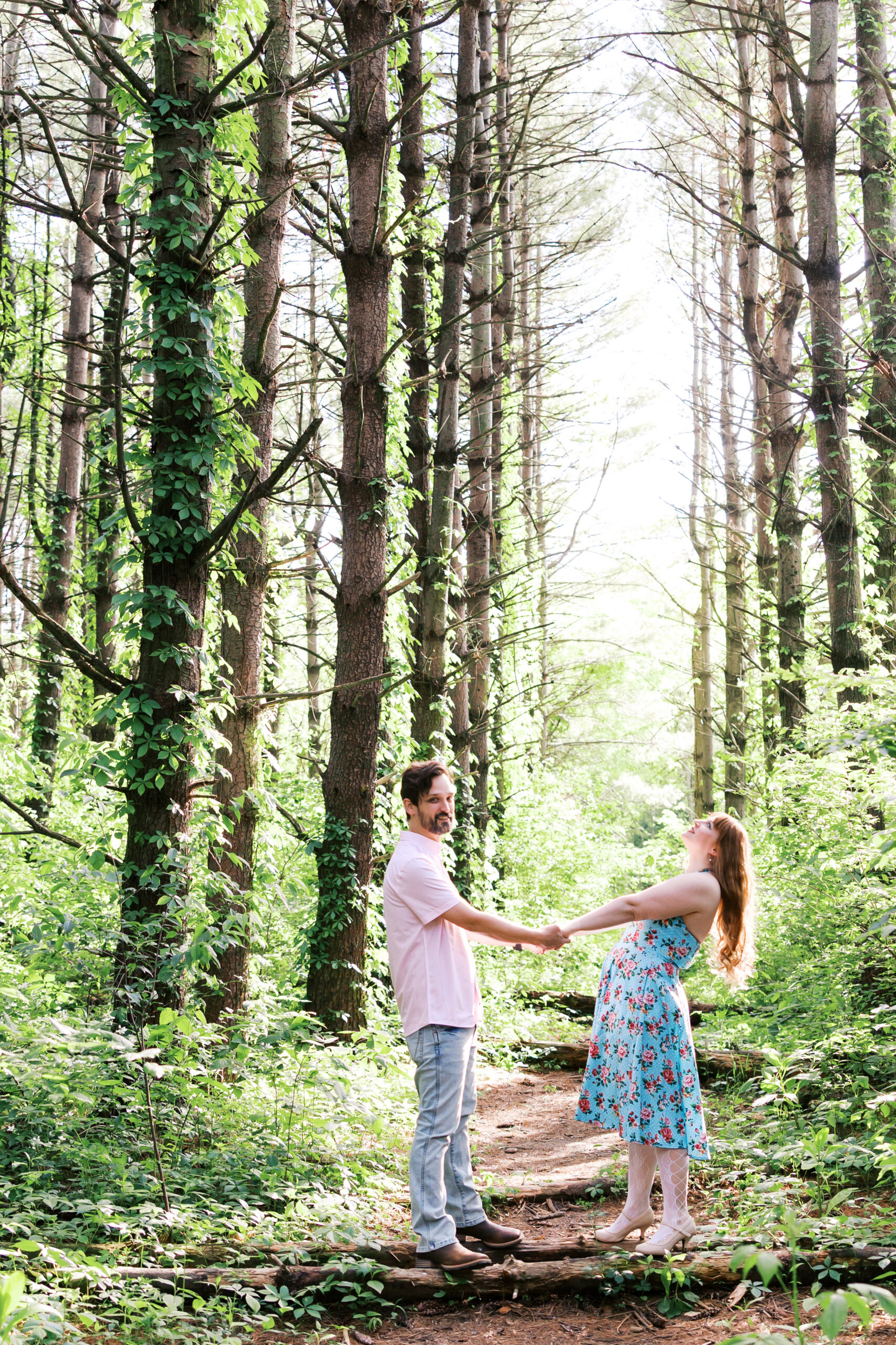Engaged couple holding hands on a wooded trail at Burr Oak, sunlight filtering through the trees."