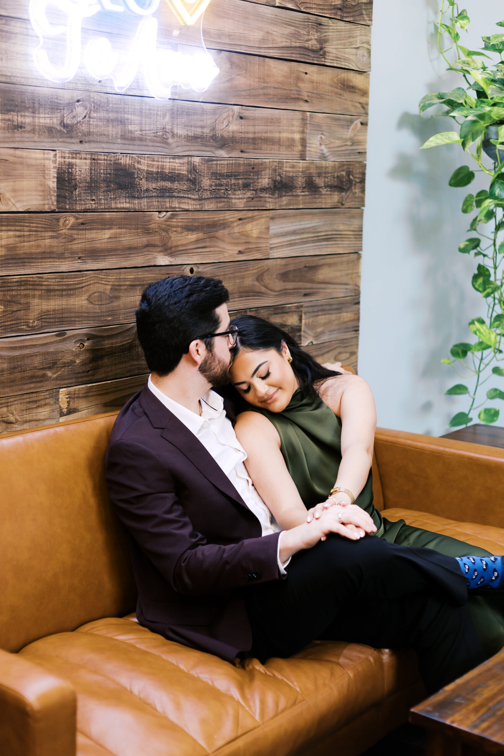 Engaged couple cuddling on a leather booth at Café Corazón, resting against a wood-plank wall.