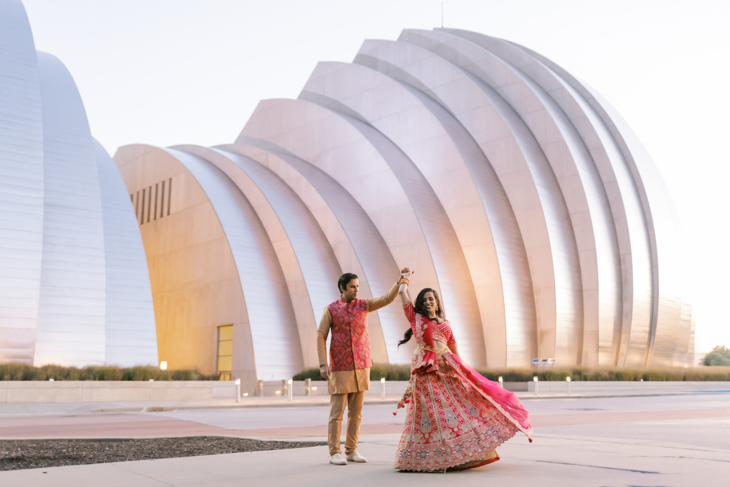 Engaged man twirling his partner in a pink lehenga, her skirt caught mid-spin, beneath the sweeping steel shells of the Kauffman Center.