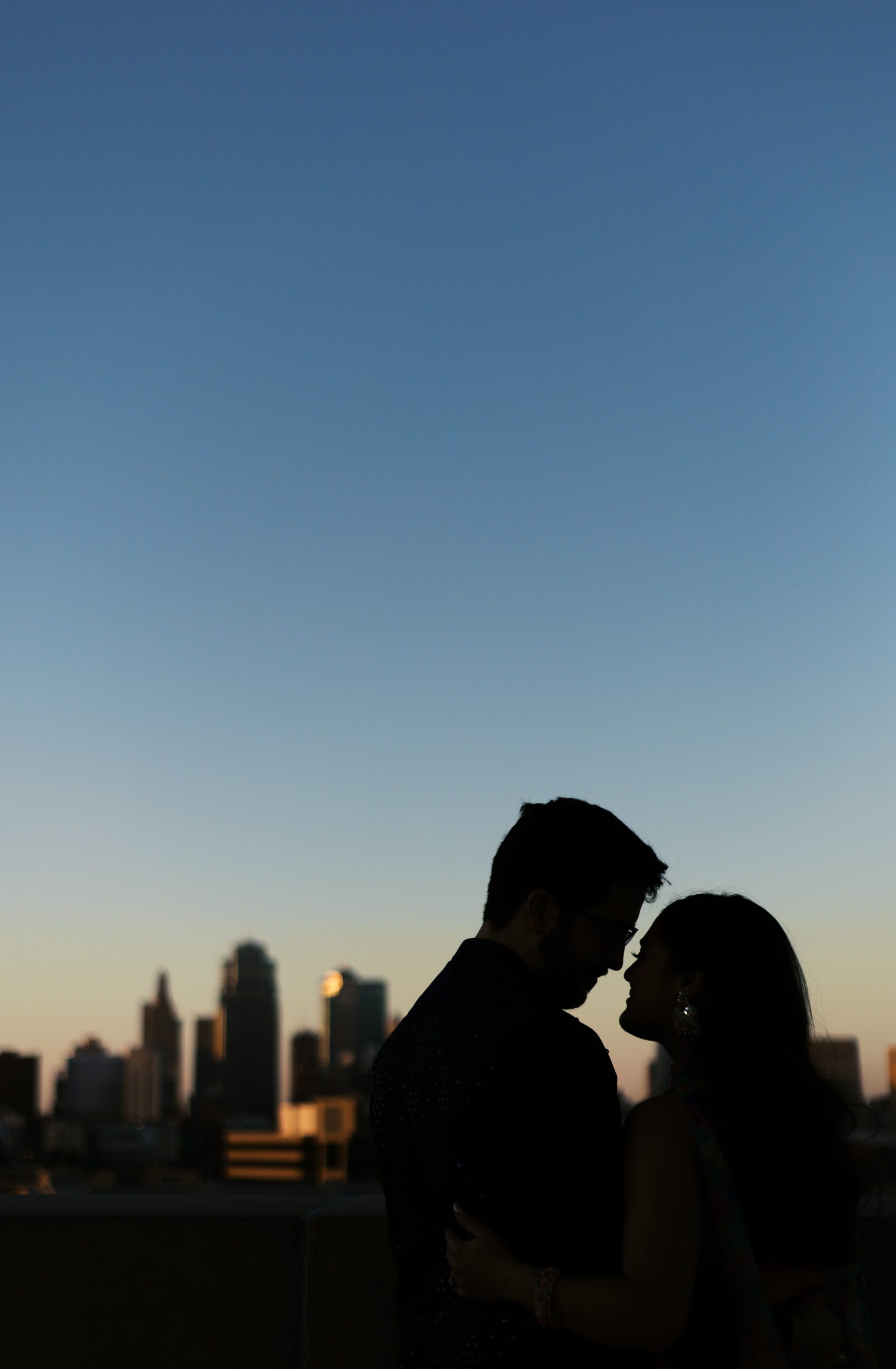 Silhouette of an engaged couple leaning in close at dusk, city skyline glowing behind them.