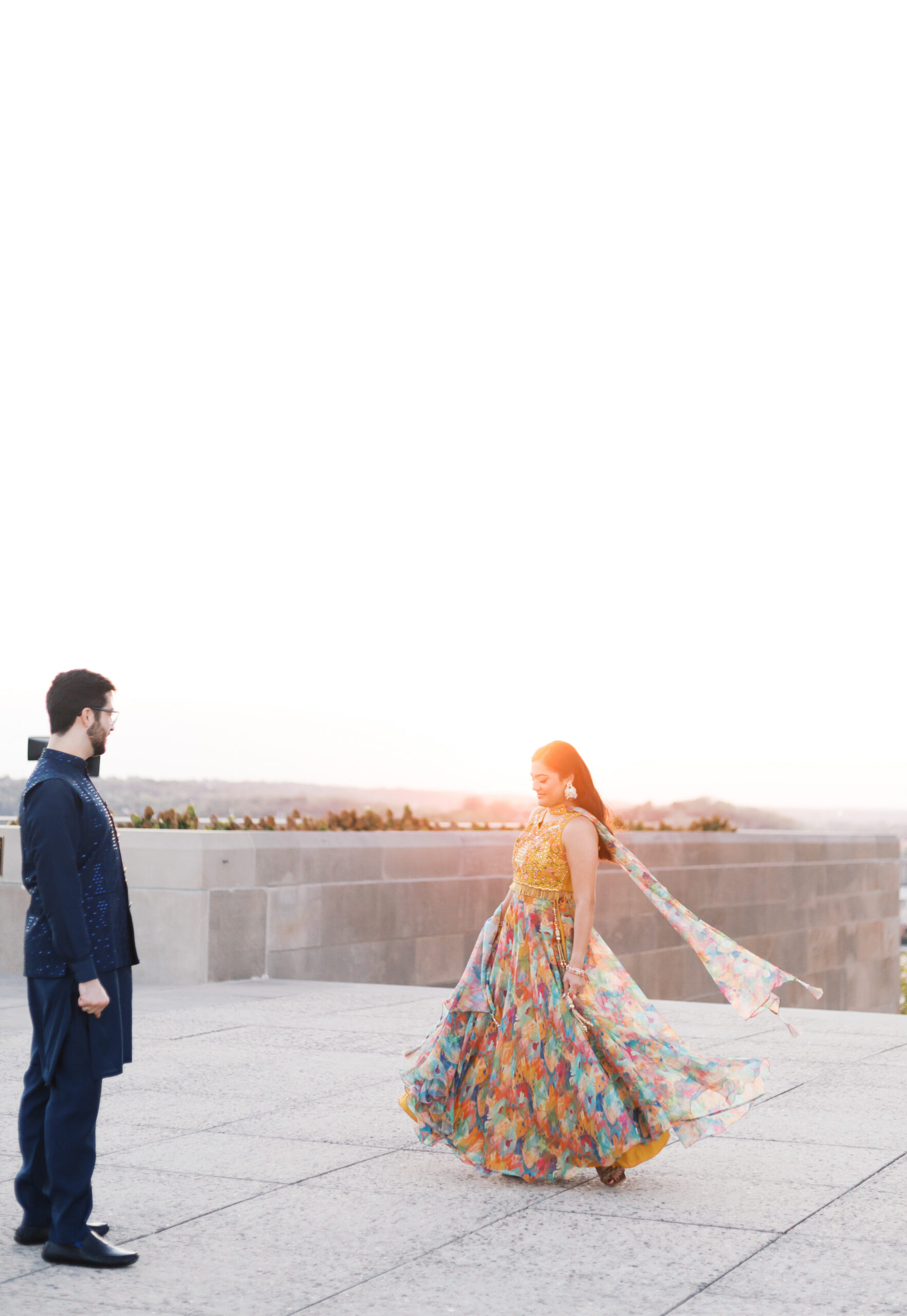 Engaged woman twirling in a colorful printed lehenga as her partner watches, golden hour light behind her.