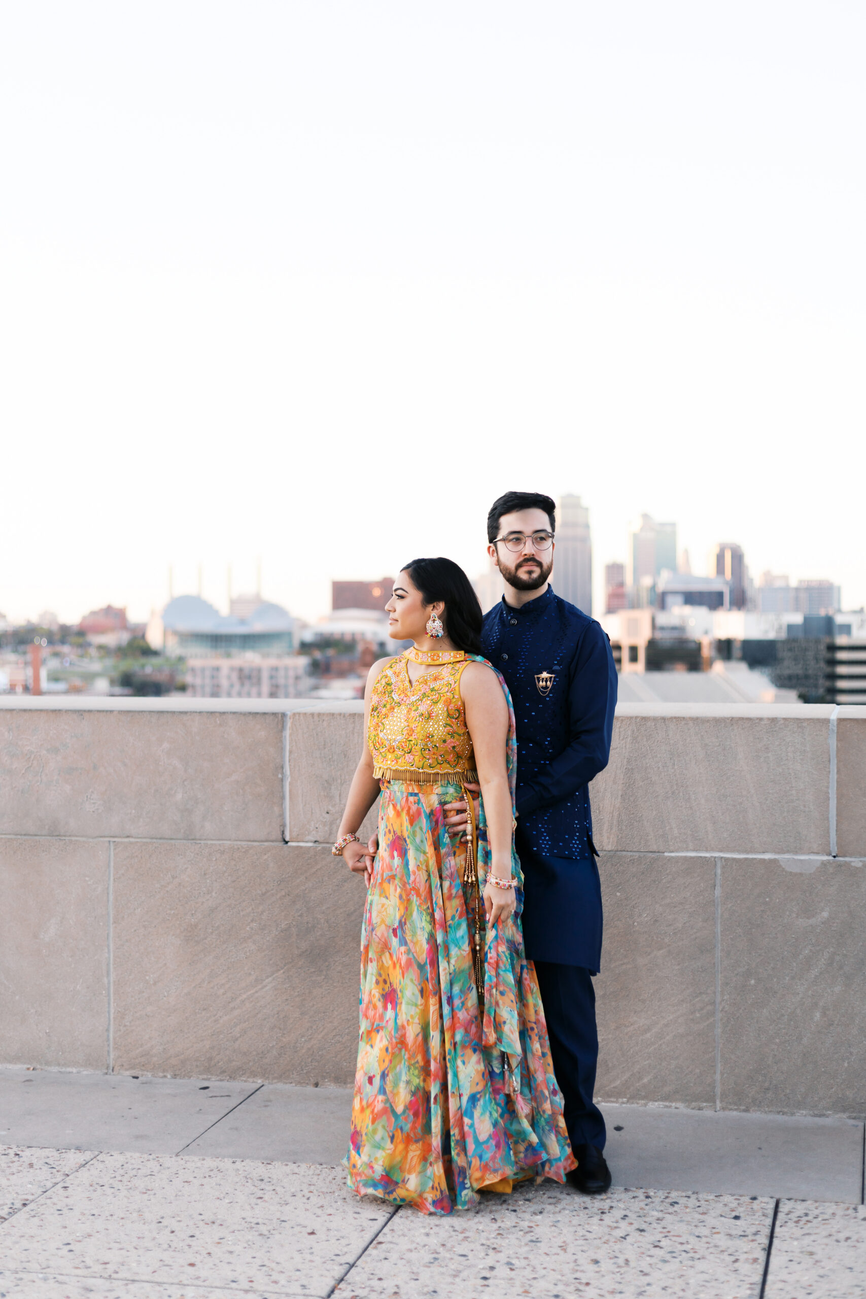 Engaged couple standing close together on a rooftop terrace, city skyline stretching out behind them.