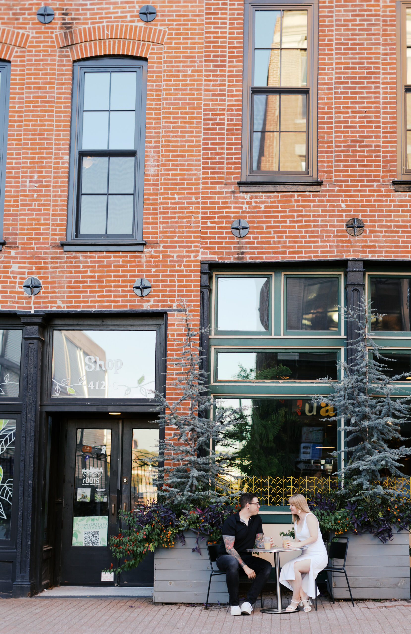 couple sitting at a café table between potted evergreens outside a brick storefront during their engagement photos.
