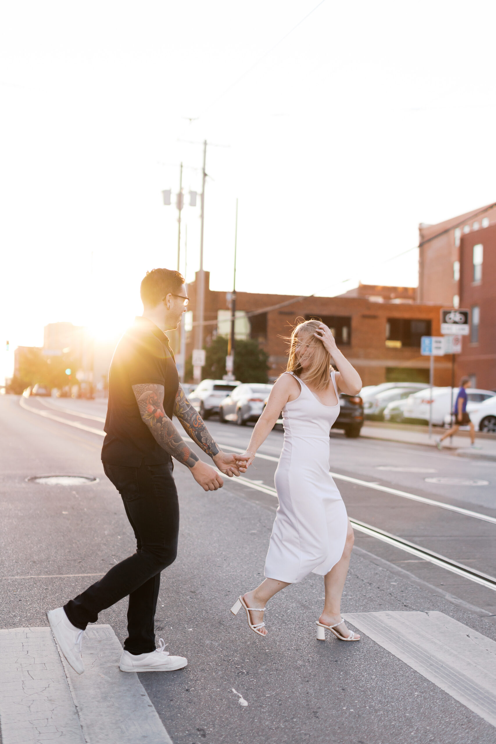 Engaged couple crossing a street hand in hand, backlit by golden hour sun.