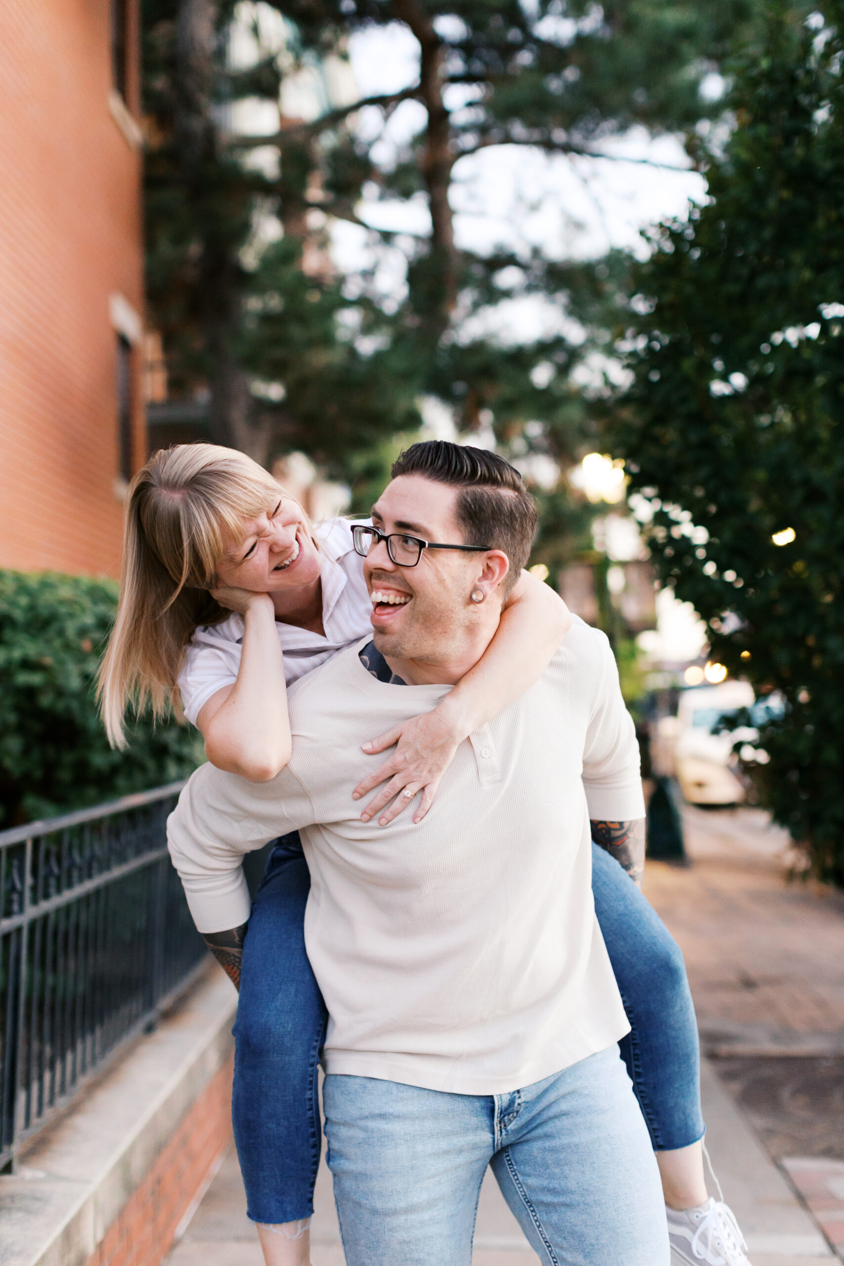 Woman laughing on her partner's back during a piggyback ride on a tree-lined sidewalk during their engagement session in Kansas City.