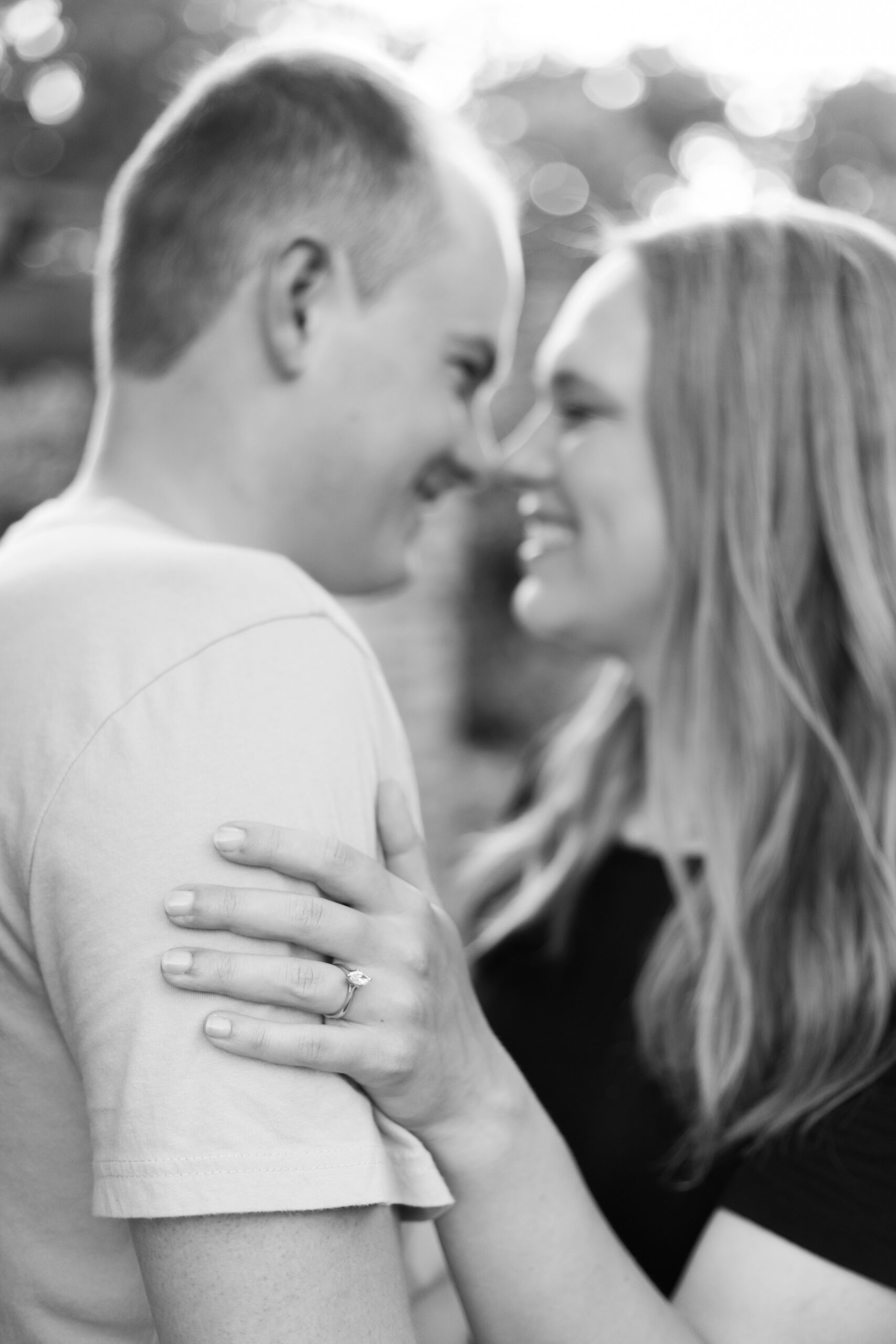 Black and white close-up of an engaged couple laughing together, her engagement ring visible on his shoulder.