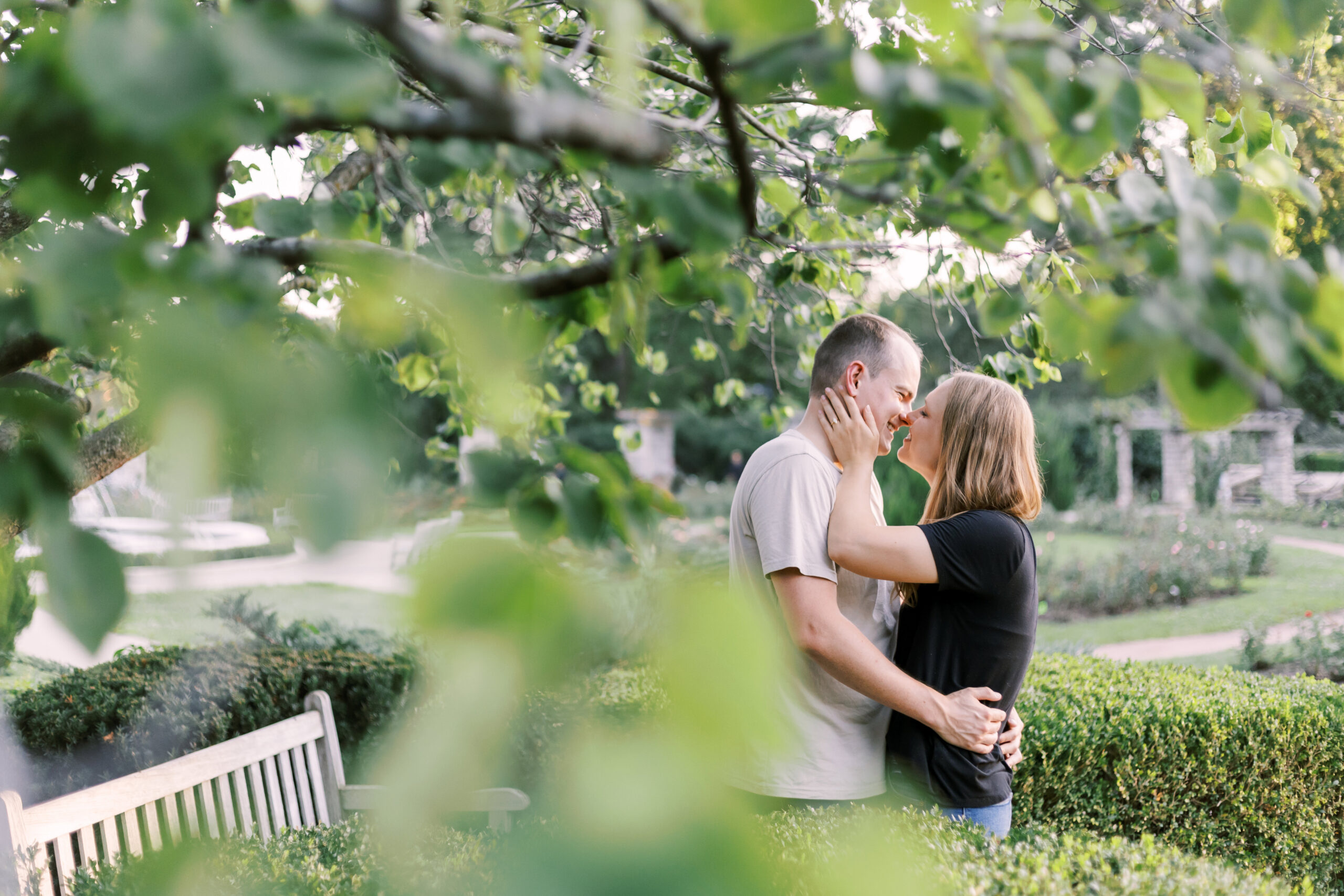 Engaged couple embracing in a garden, glimpsed through leafy branches in the foreground.