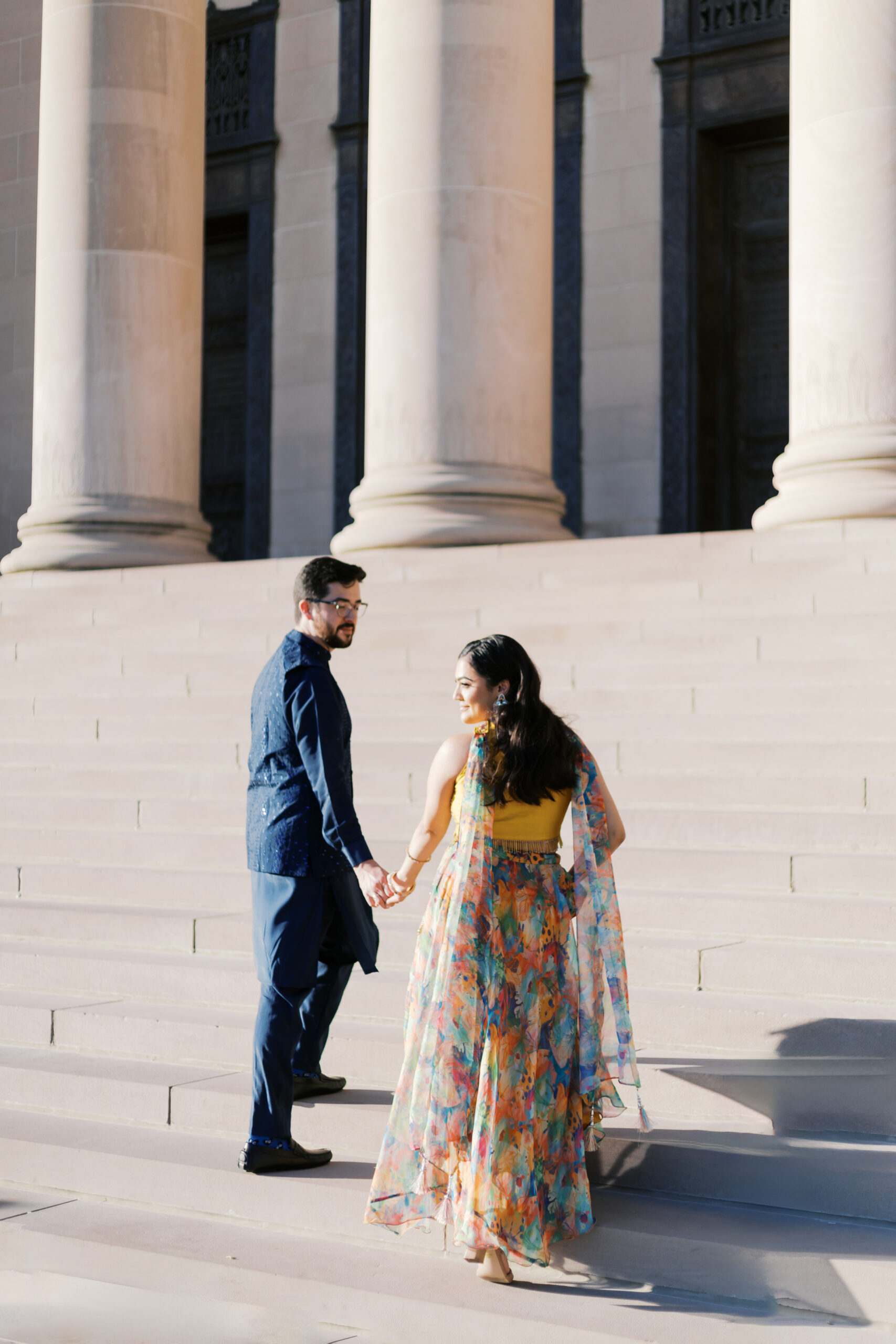 "Engaged couple walking hand in hand up grand stone steps between towering columns.