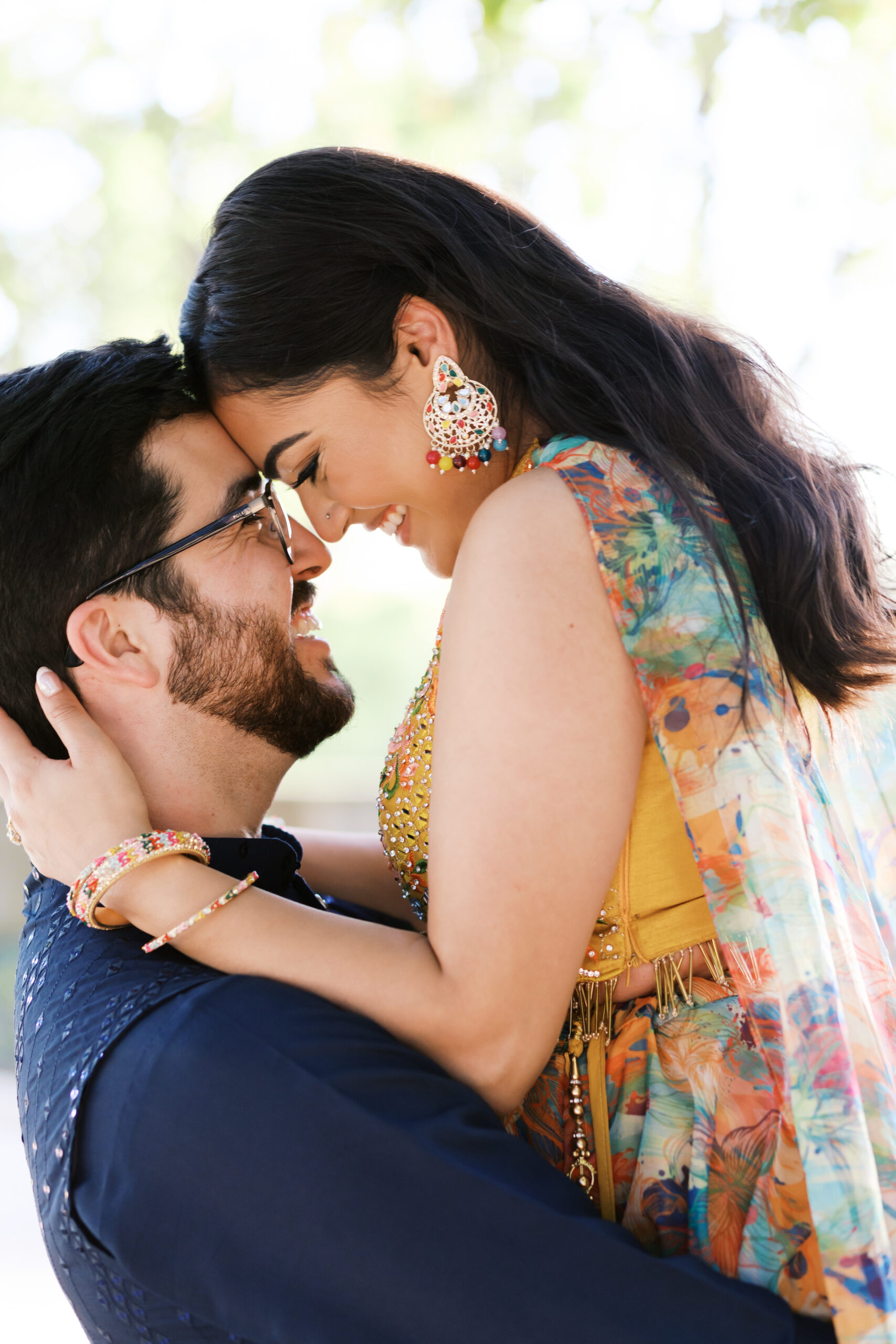 Close-up of an engaged couple leaning forehead to forehead, laughing together in dappled sunlight.