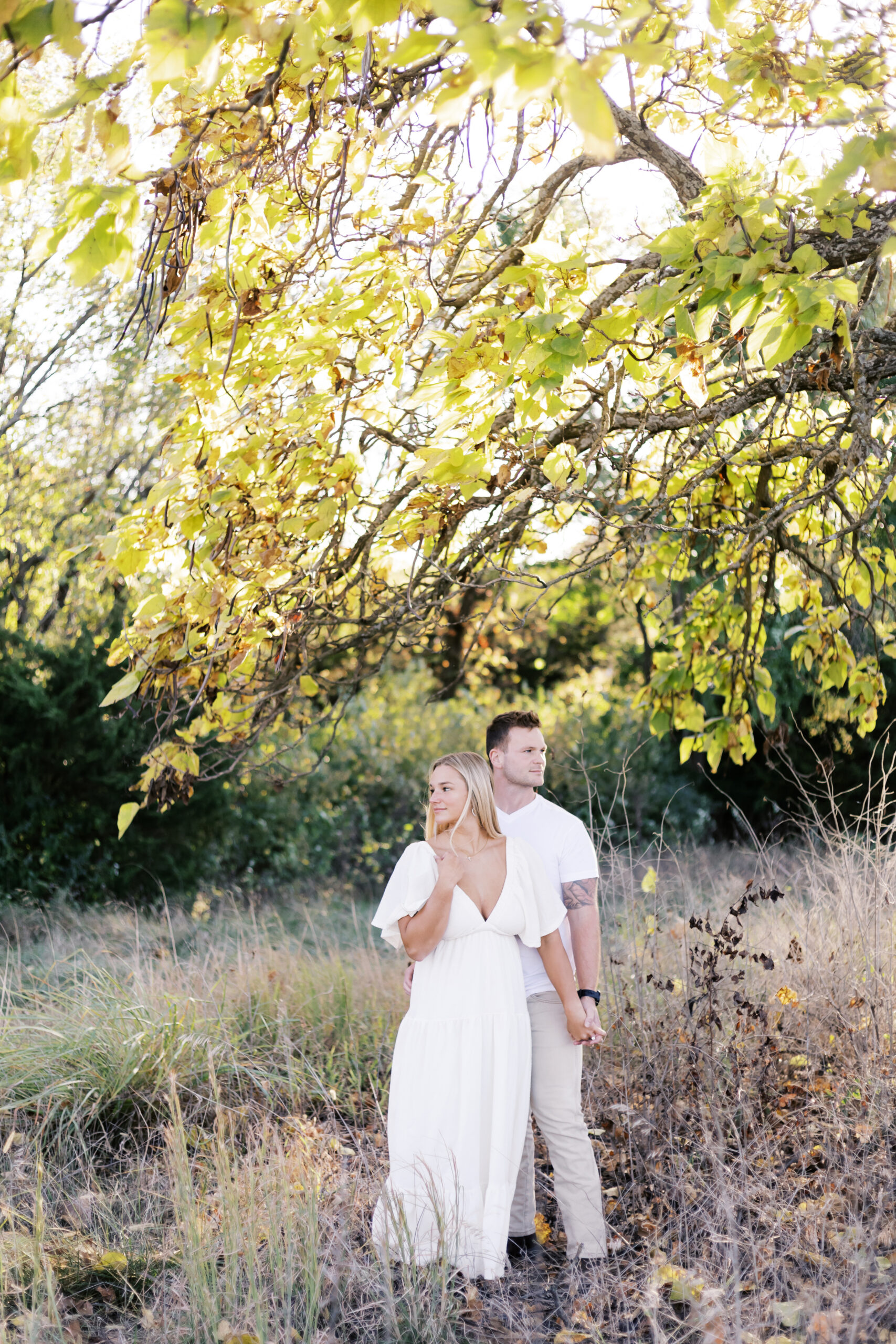 Engaged couple holding hands beneath golden leaves in a wooded clearing