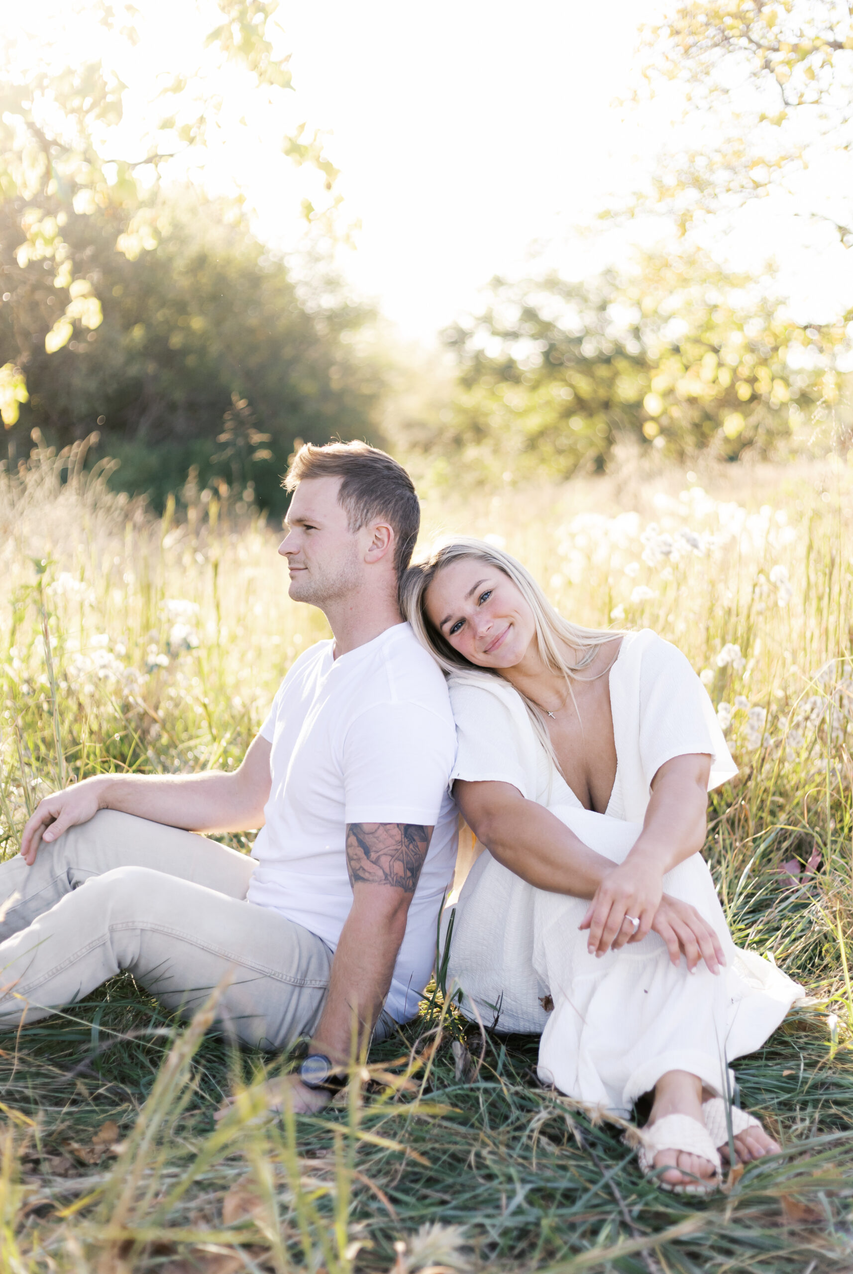 Engaged couple sitting back to back in a field of wildflowers, smiling at the camera