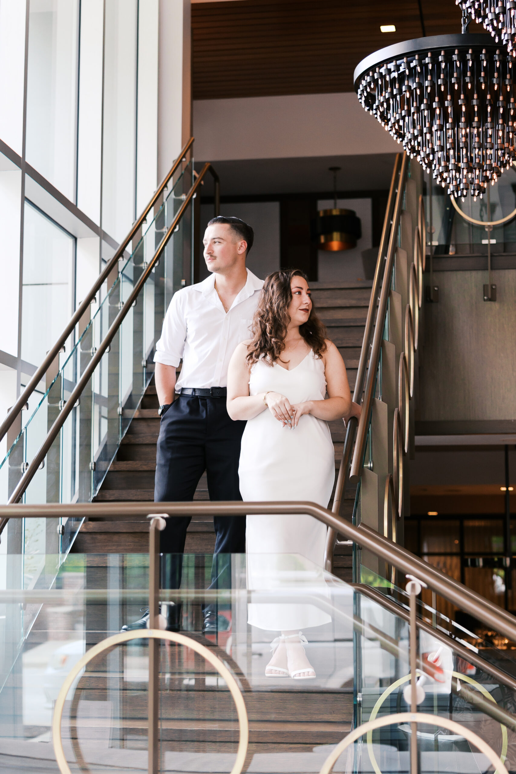 Couple standing on a staircase beneath a dramatic pendant chandelier during their engagement session at Stockhill