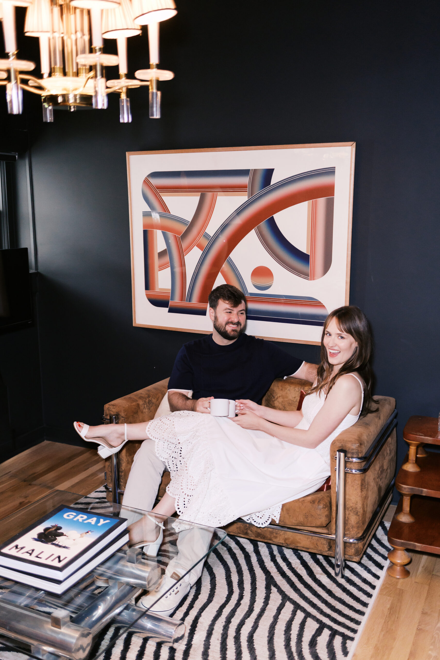 Engaged couple sitting together in a leather armchair, coffee mugs in hand, beneath a bold abstract painting.