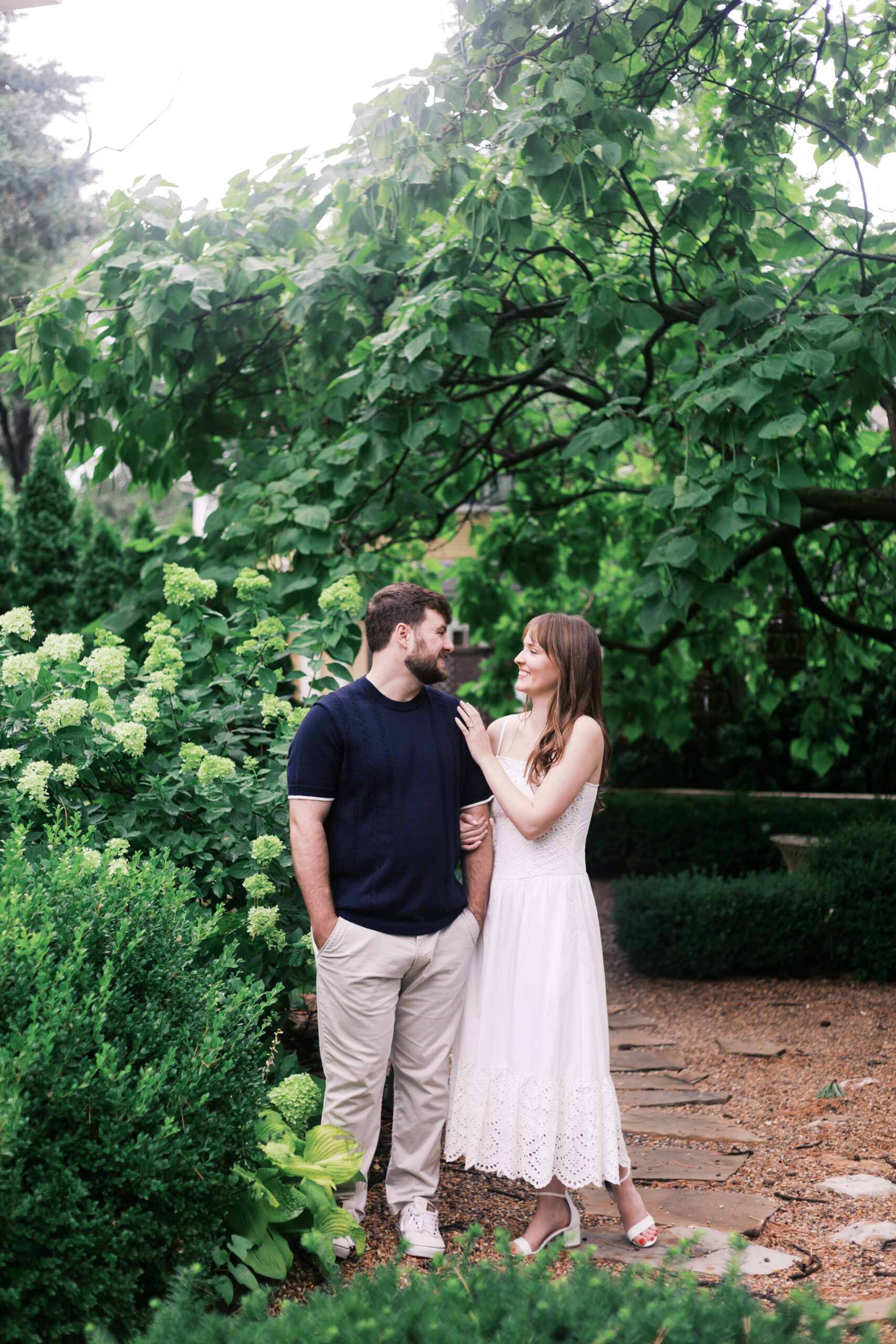 Engaged couple standing close under a canopy of green leaves, surrounded by hydrangeas outside the Truitt in Kansas City.