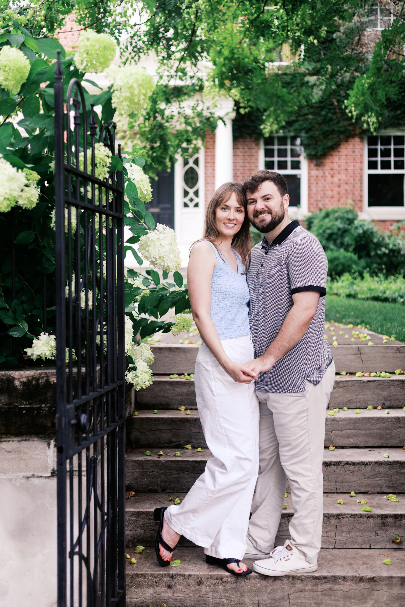Couple holding hands on brick steps beside a hydrangea-covered gate during their Kansas City engagement session