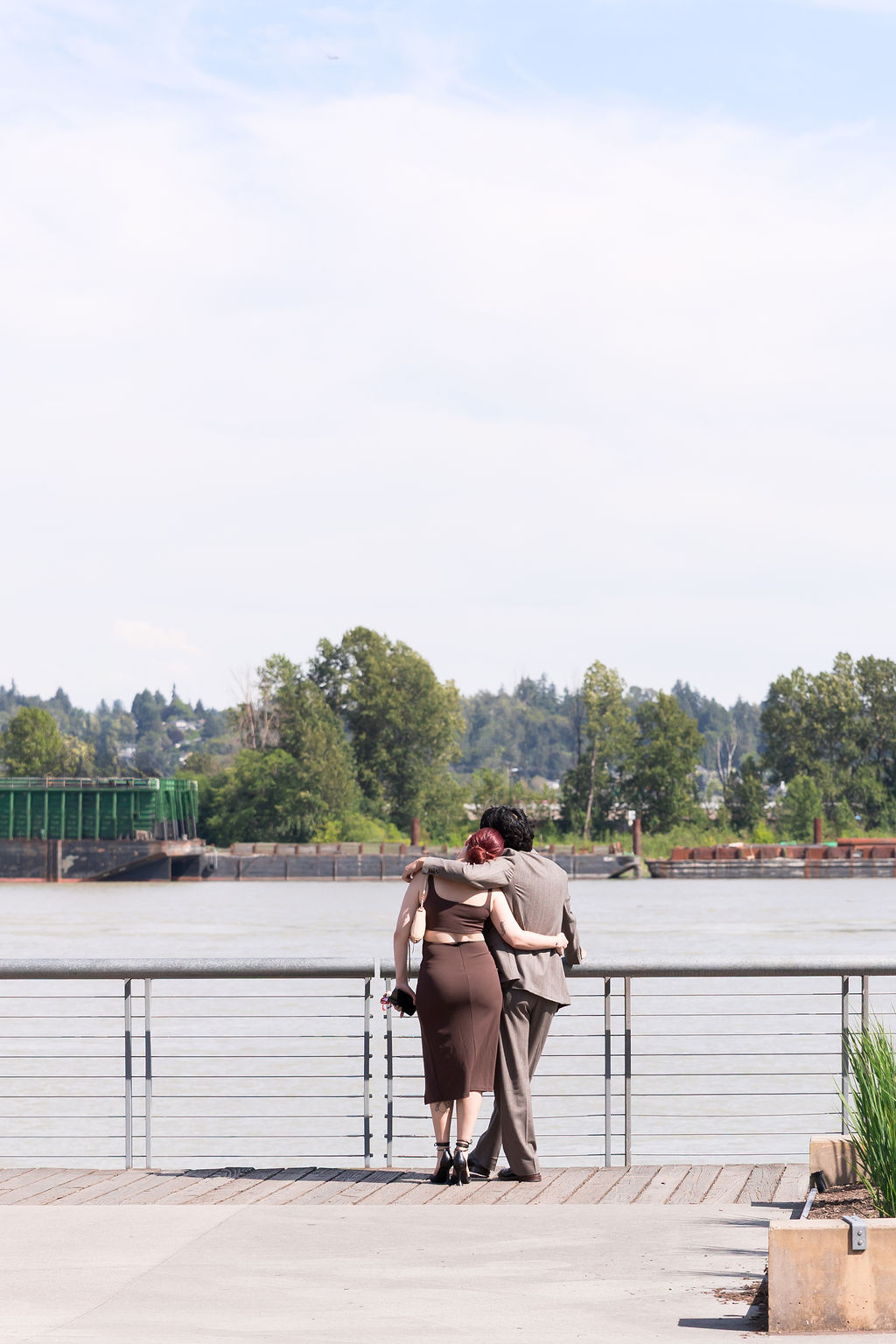 Couple stands with their arms around each other, looking out over the river from a waterfront boardwalk.