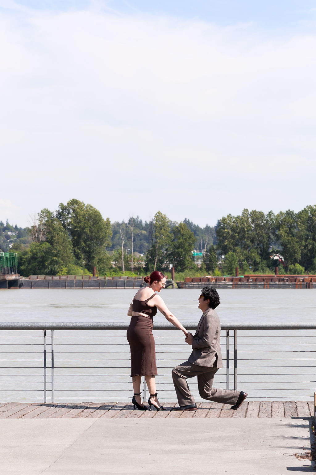 One partner kneels while holding the other's hand during a surprise proposal by the river.