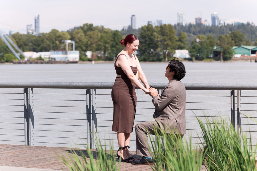Close view of the proposal as one partner kneels and holds both hands on a sunny waterfront.