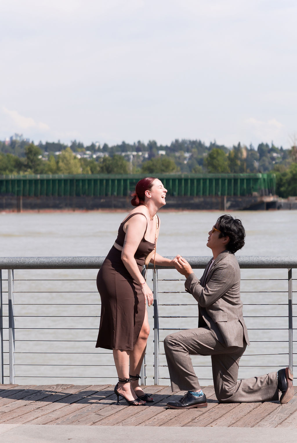 The person being proposed to laughs with surprise as their partner kneels and holds their hand.