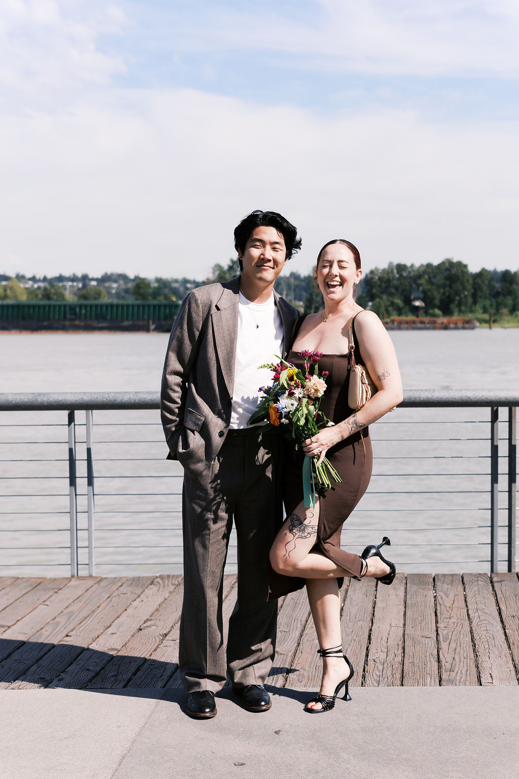 The engaged couple poses together with flowers on a waterfront boardwalk.