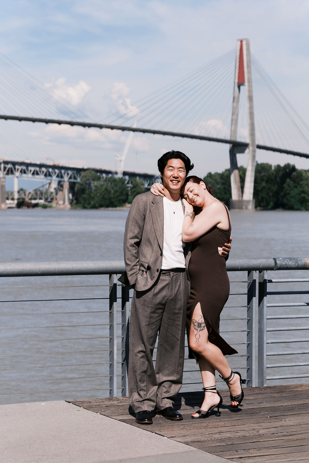 The engaged couple stands together with a bridge and river in the background.