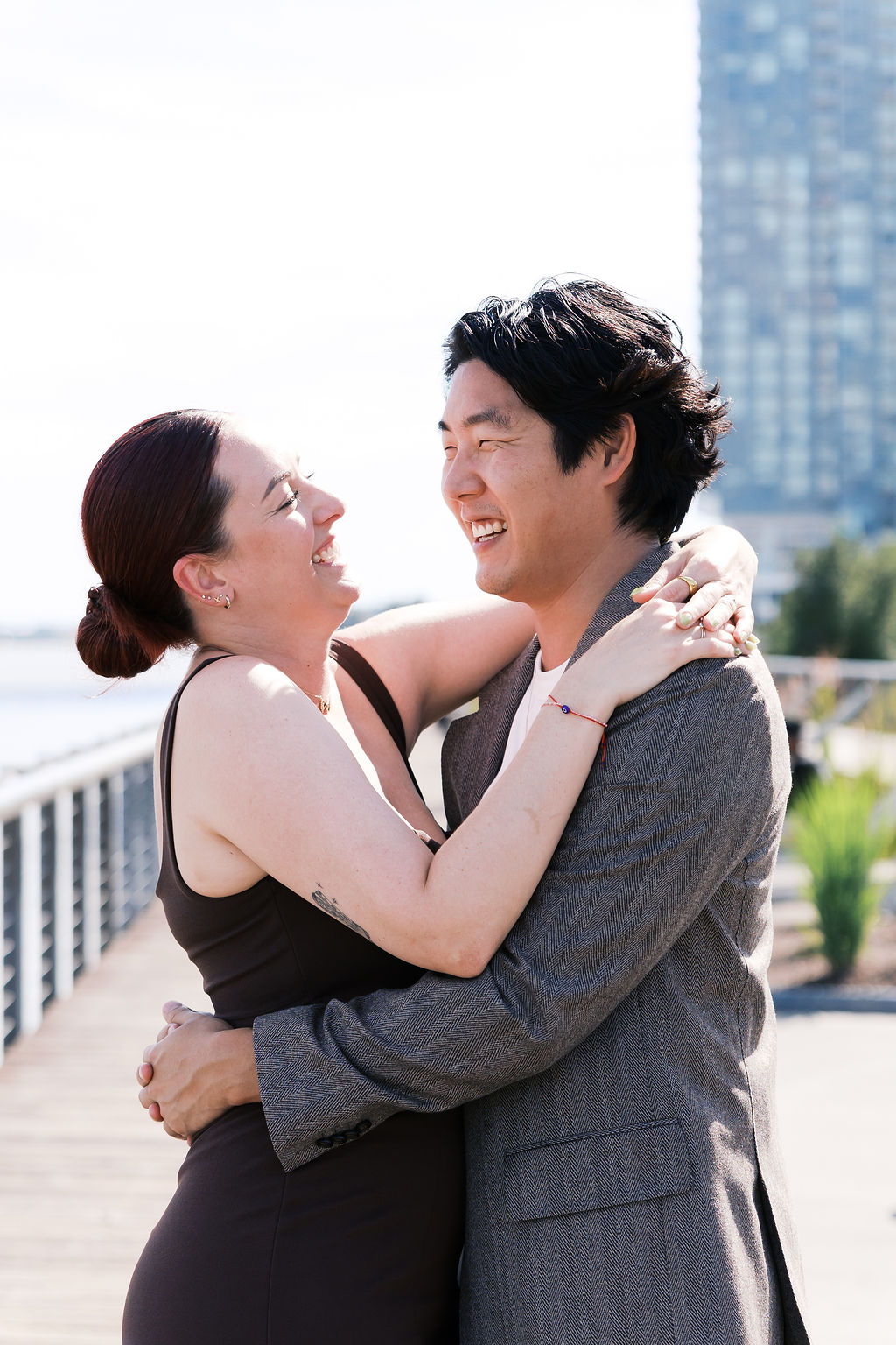 The couple laughs together while embracing on a sunny riverside boardwalk.