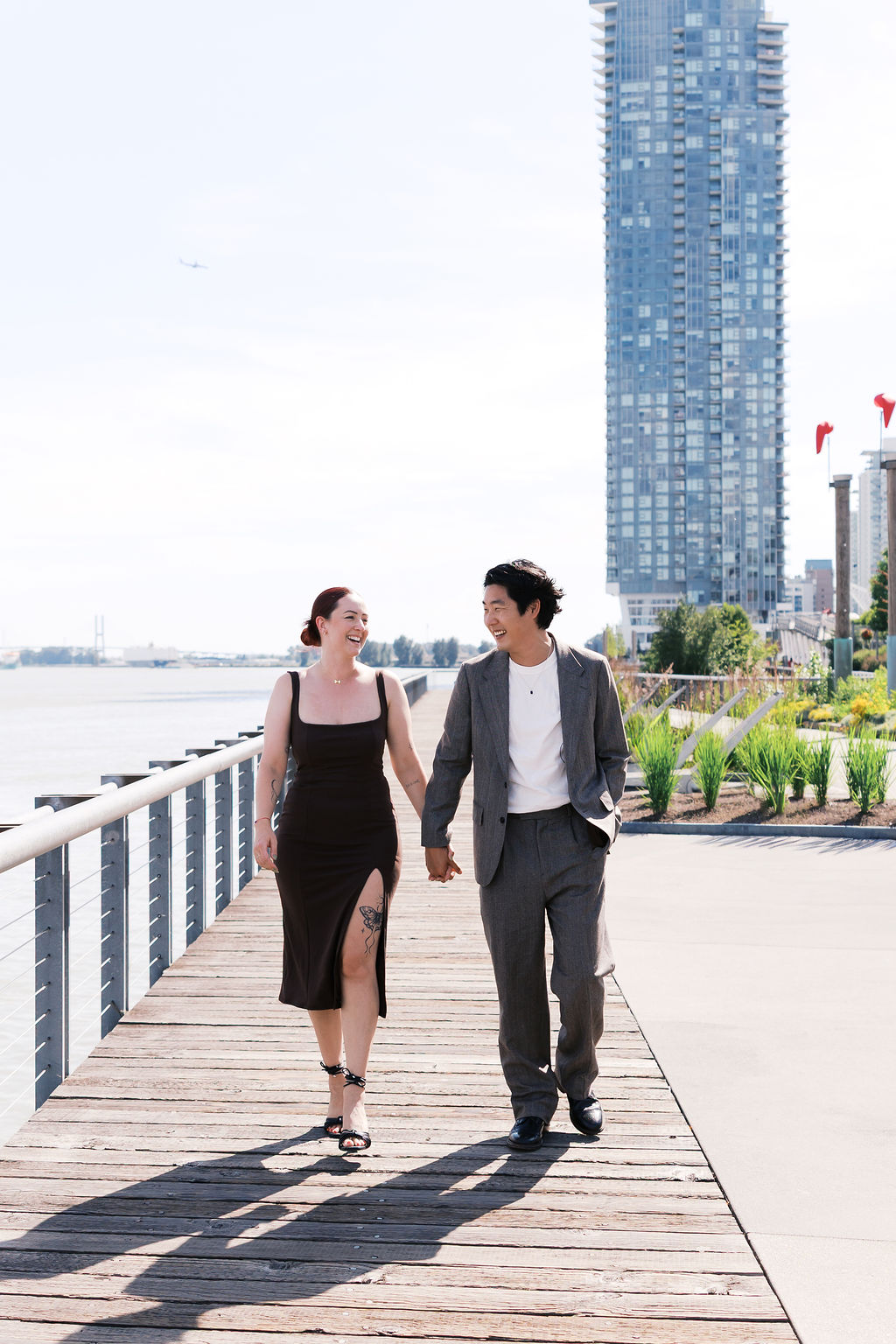 Newly engaged couple walks hand in hand along a sunny waterfront boardwalk, smiling at each other.