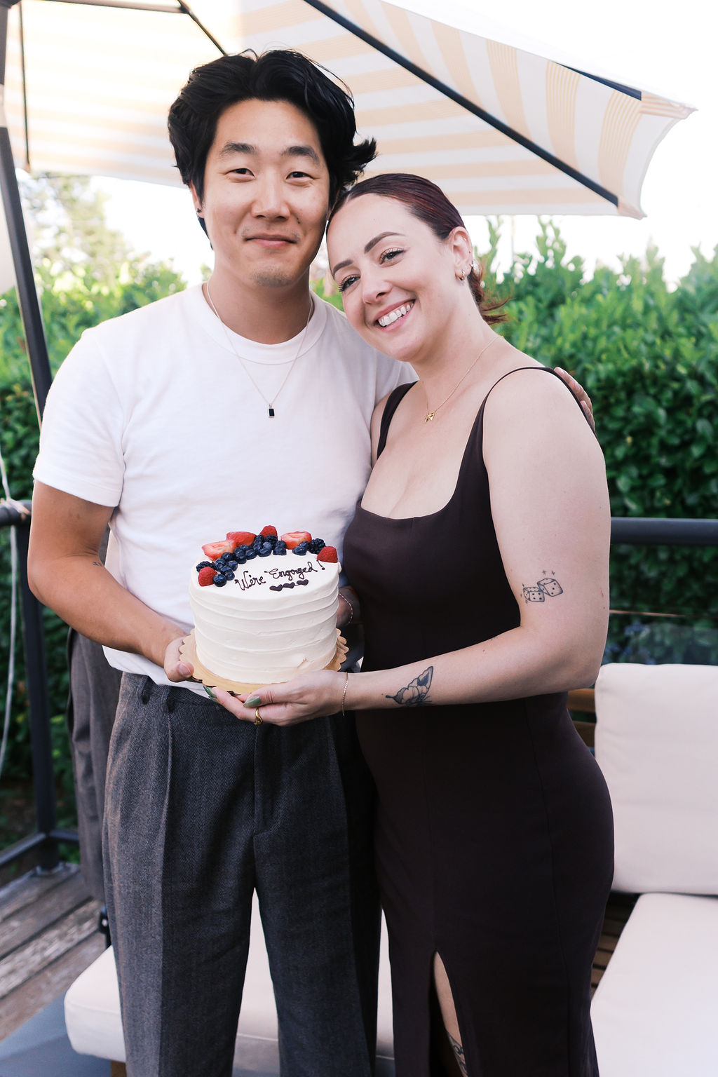 The engaged couple smiles while holding a cake decorated with fresh berries and "We're Engaged!"