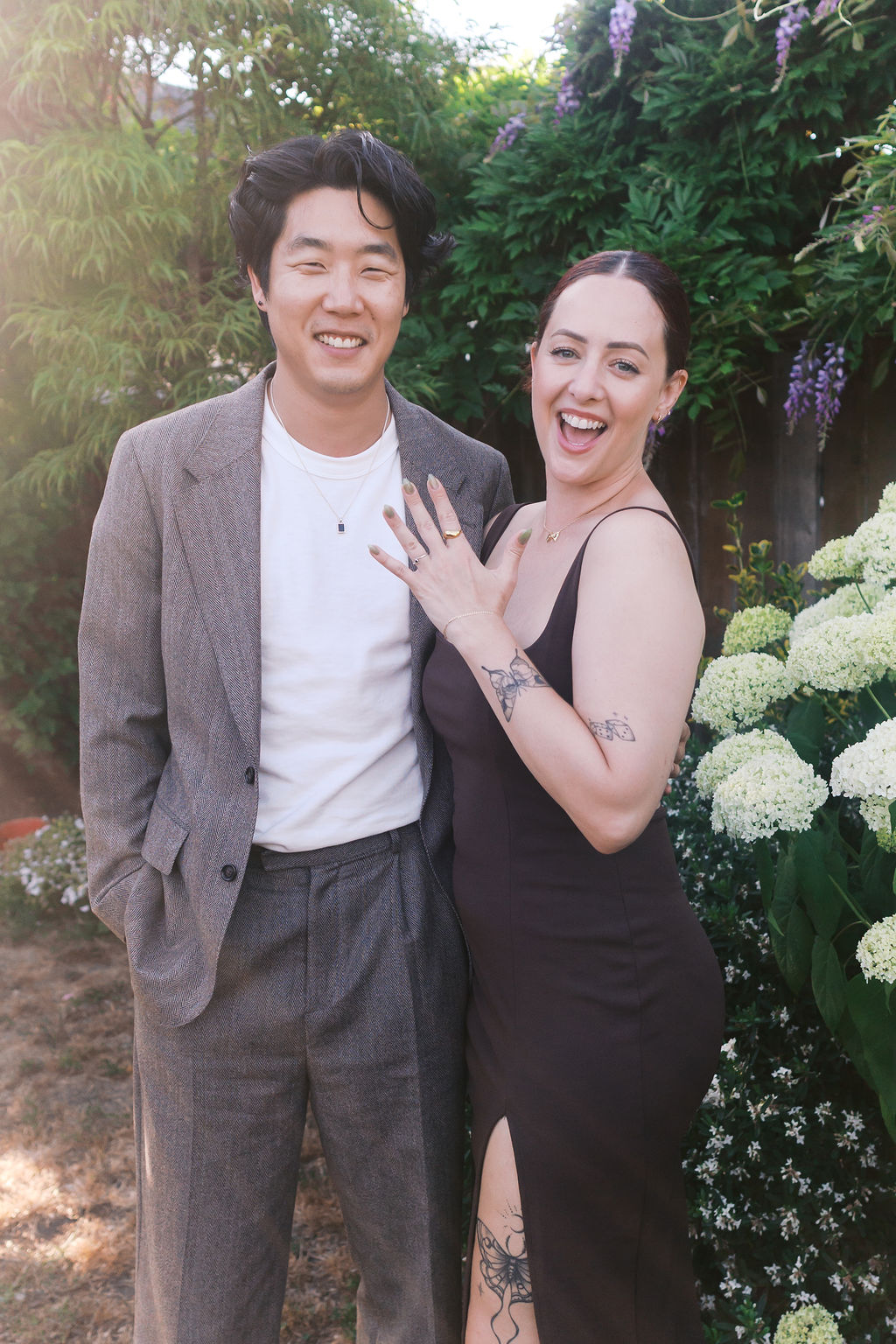 The newly engaged couple poses together in a garden, showing the engagement ring.