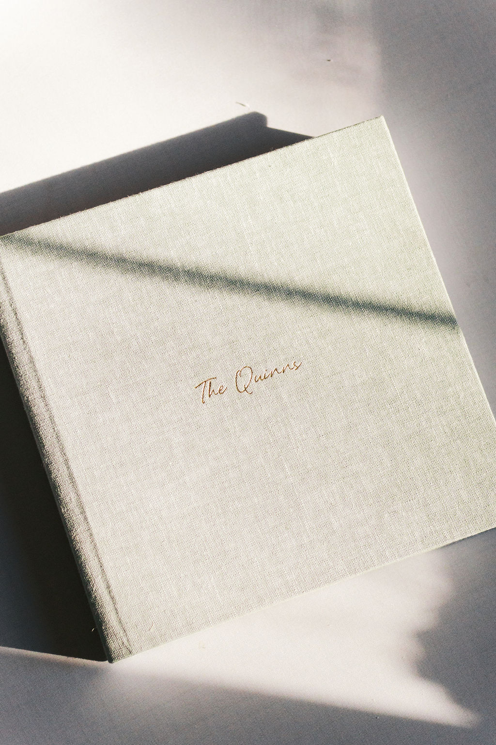 Cream linen wedding photo album with gold script lettering, resting in soft sunlight and shadow.