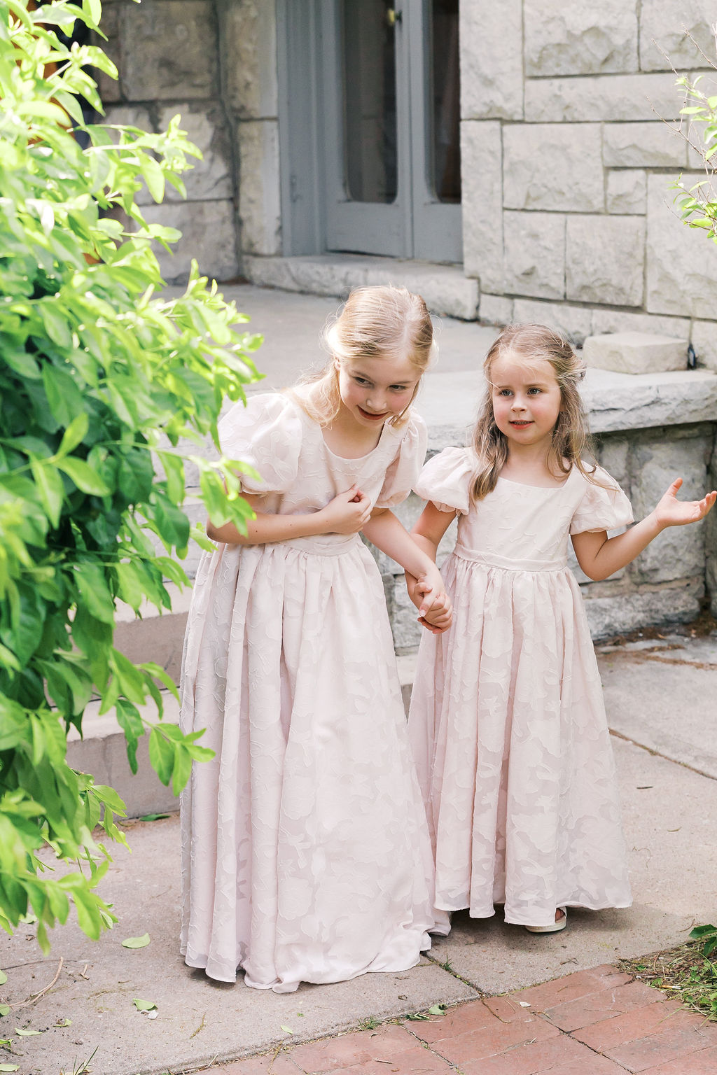 Two young flower girls in matching pale pink dresses holding hands outside a stone building.