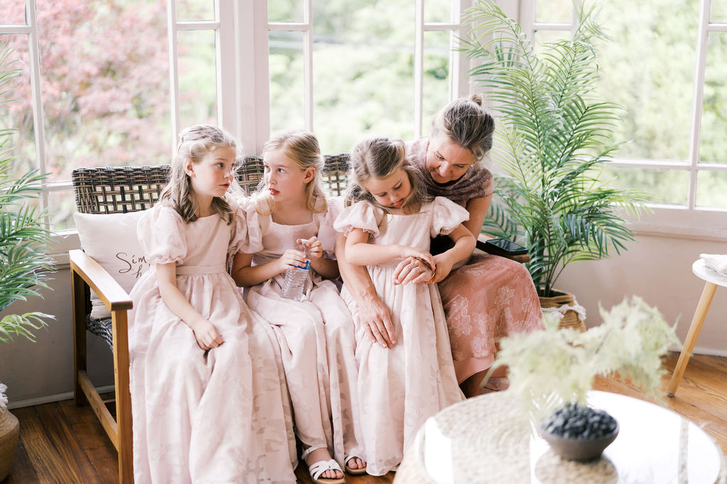 Three flower girls in matching pink dresses sitting beside a woman in a bright, plant-filled room.