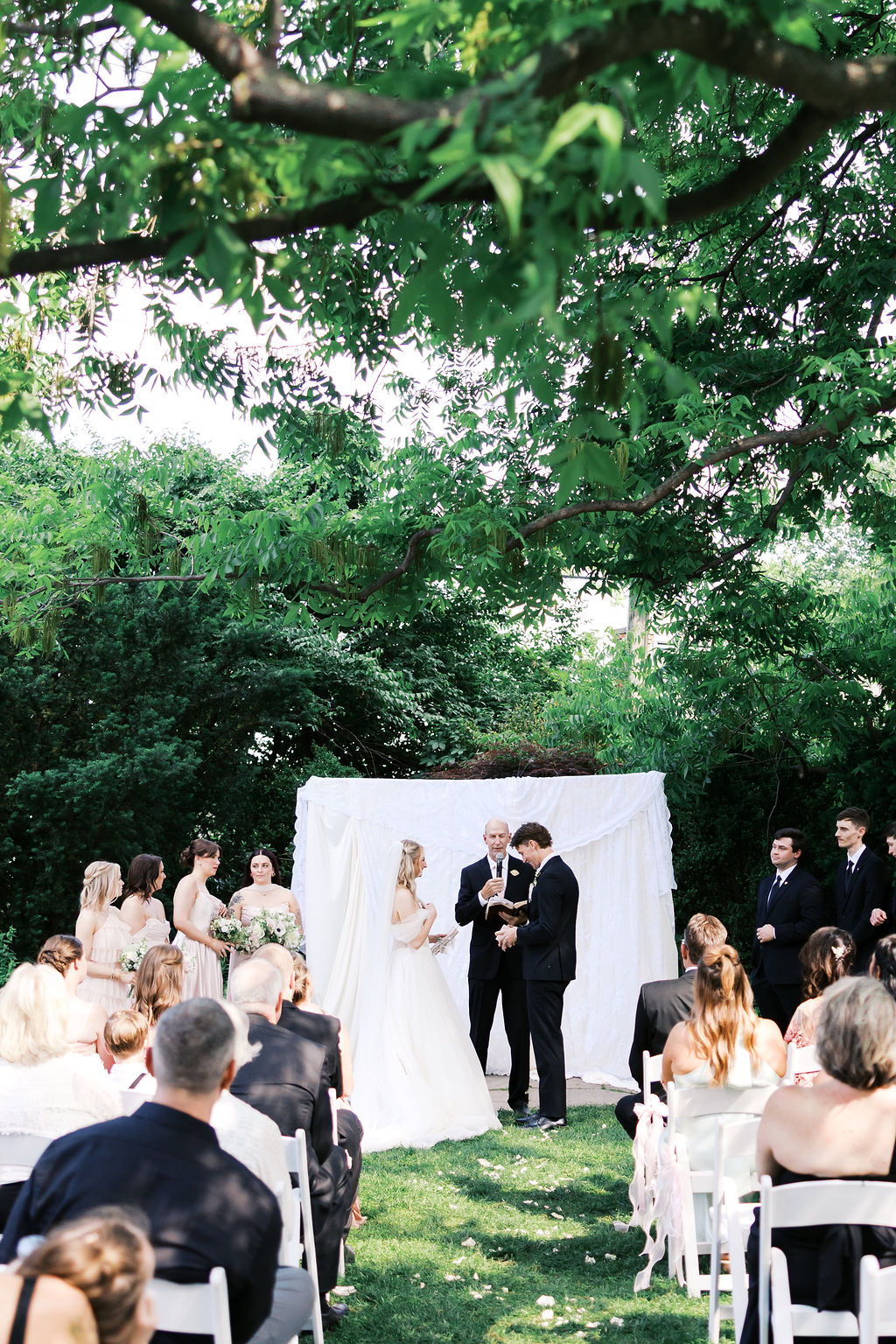 Bride and groom standing with their wedding party during an outdoor ceremony beneath leafy trees.