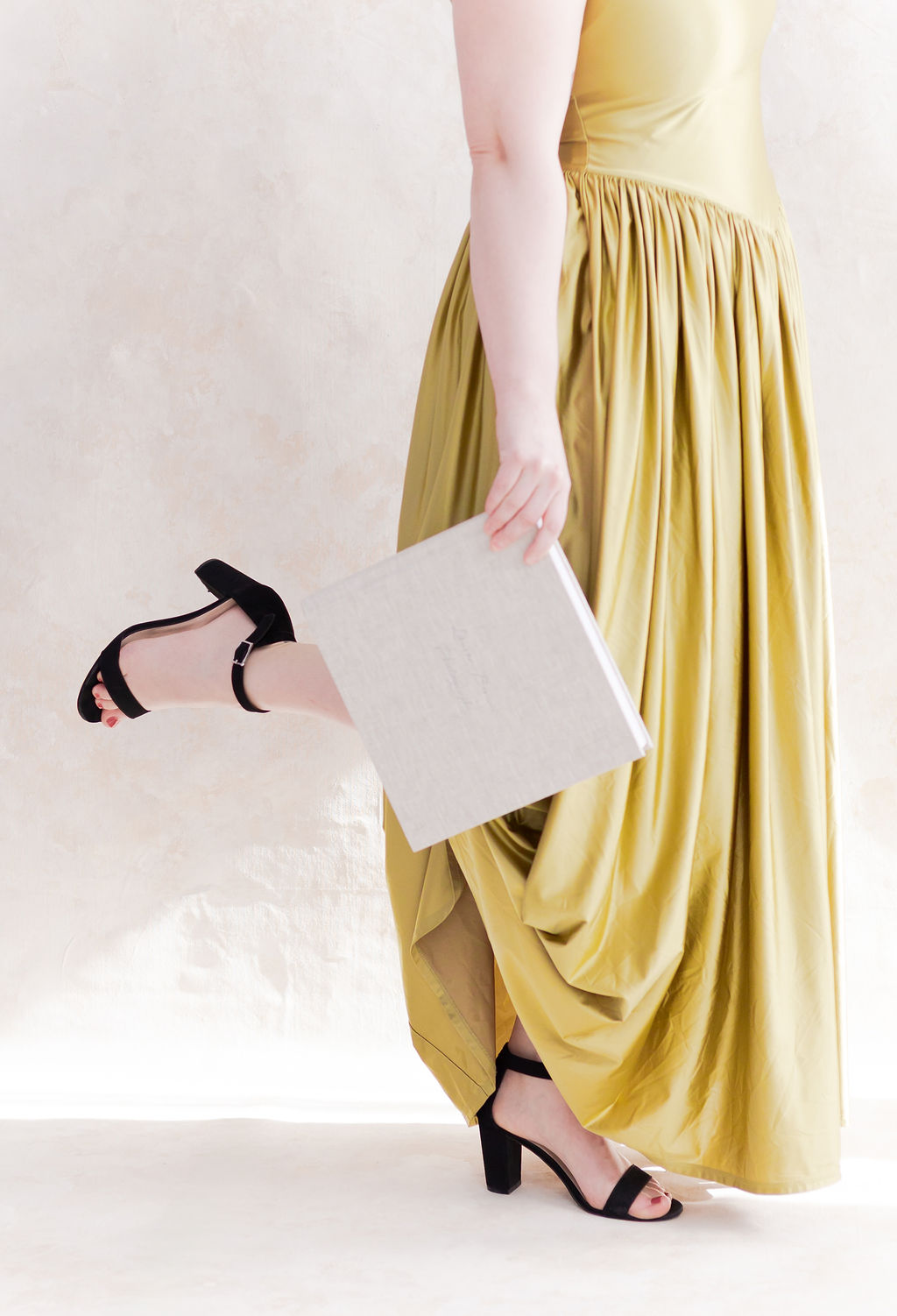 Person in a flowing olive green dress and black heels carrying a neutral linen photo album.
