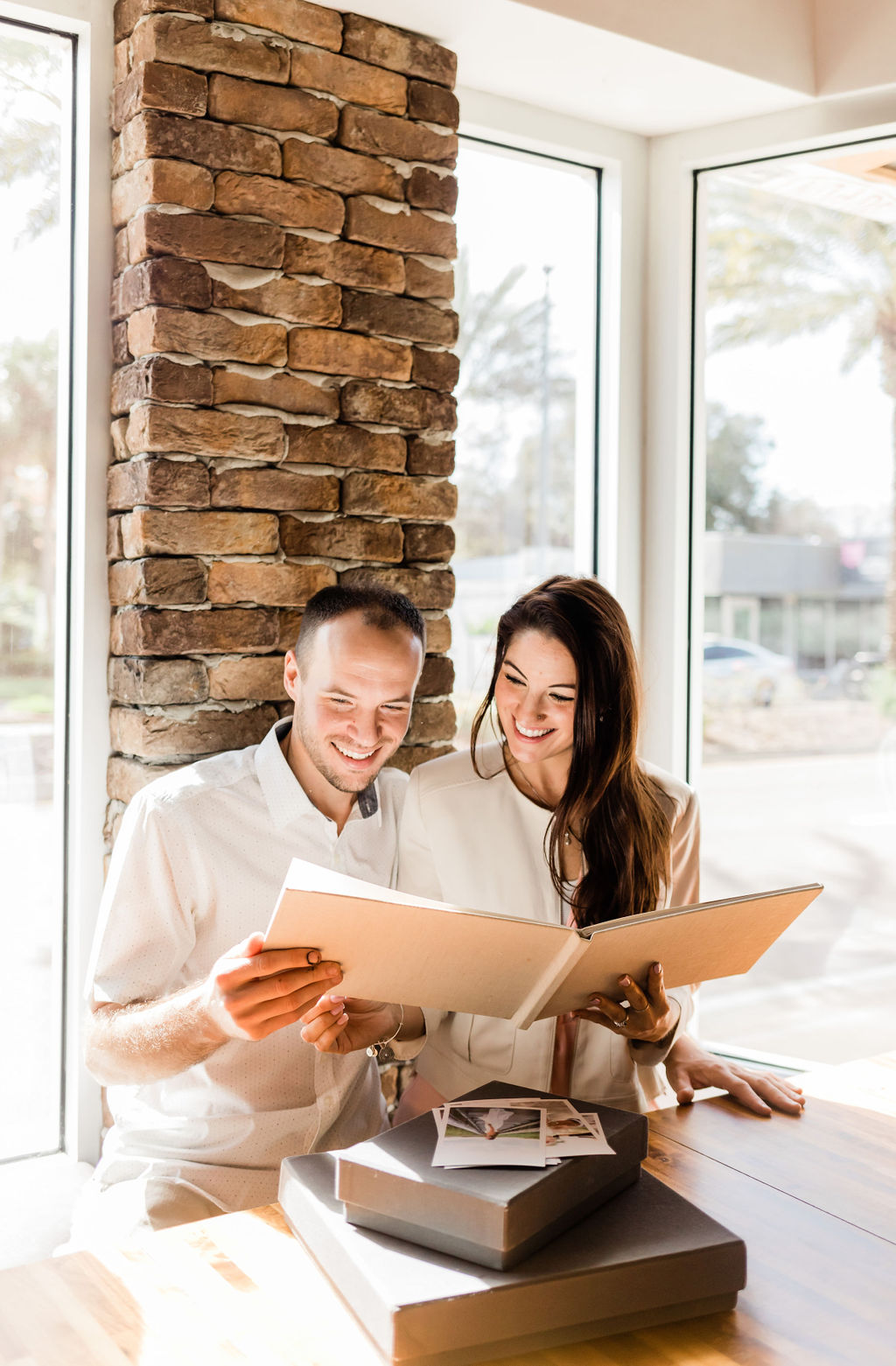 Couple smiling together while looking through a wedding album at a sunlit table.
