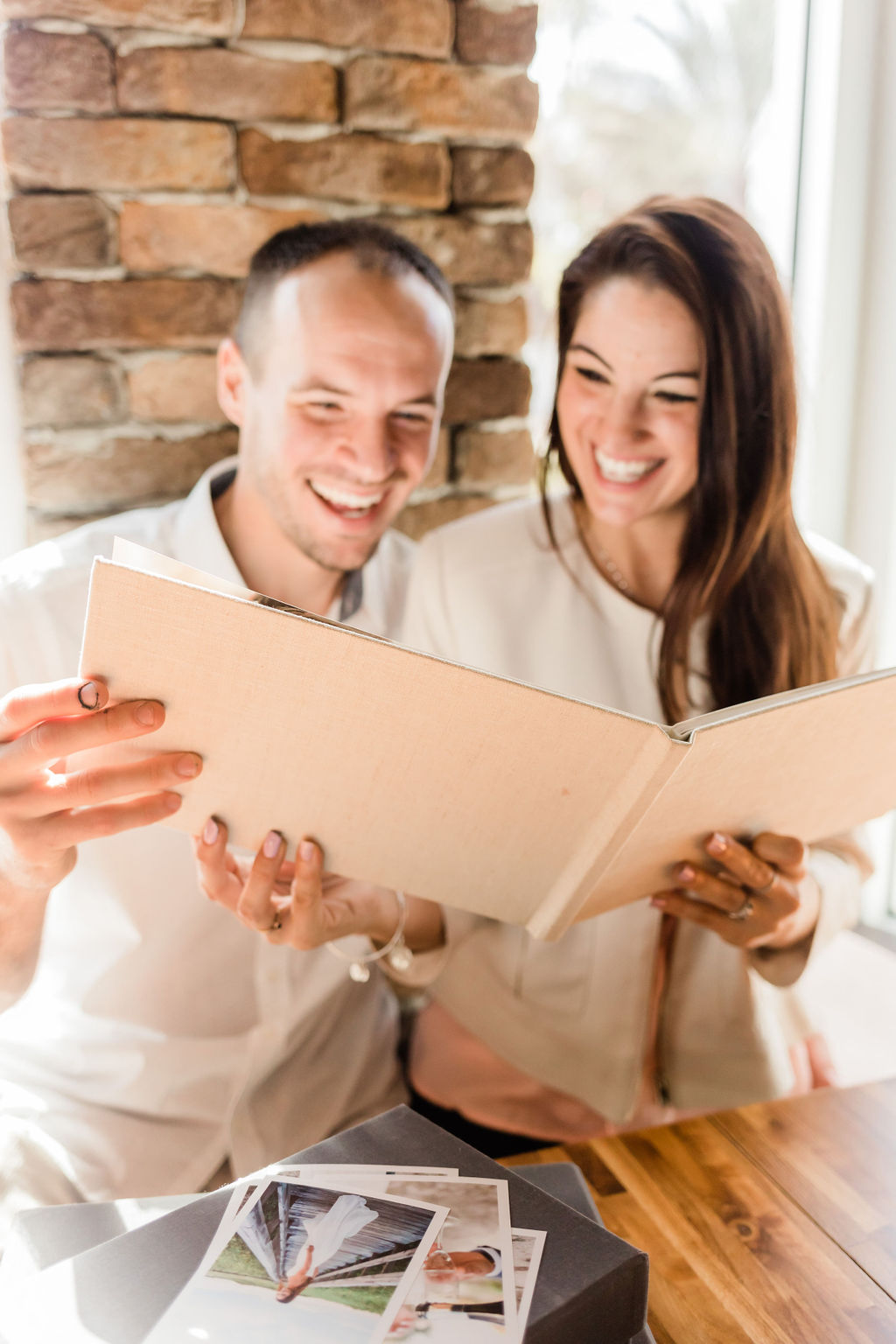 Close-up of a smiling couple looking through a linen wedding album with printed photographs on the table.
