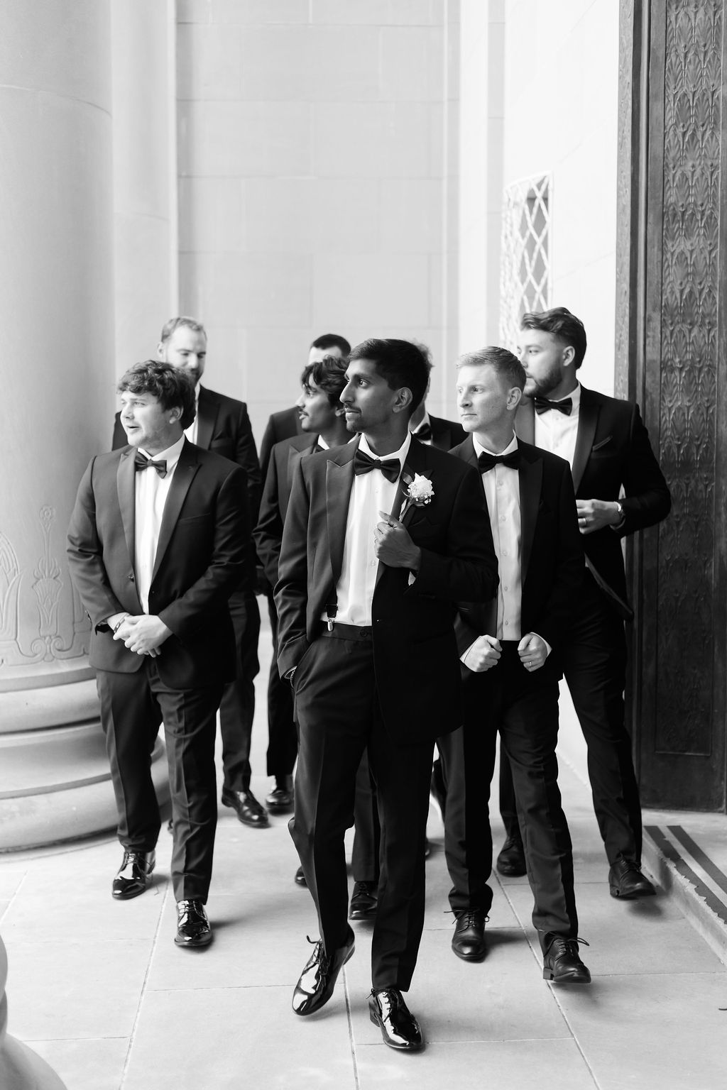 Wedding party in black tuxedos walking through a grand stone colonnade, photographed in black and white.