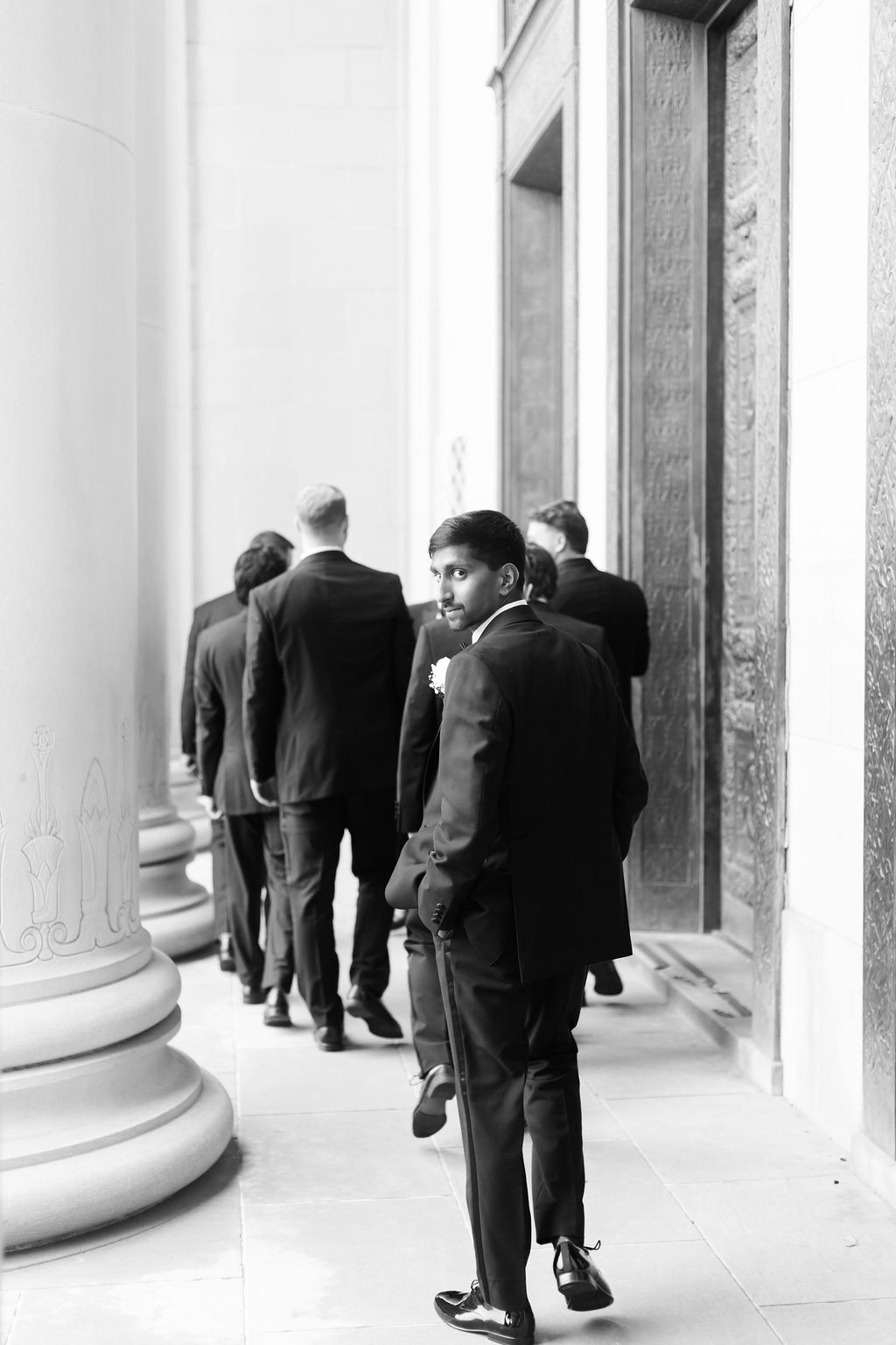 Person in a black tuxedo looks back toward the camera while walking with the wedding party through stone columns.