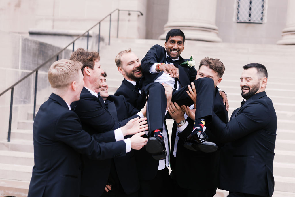 Wedding party in black tuxedos lifts a smiling person in the air outside a grand stone building.